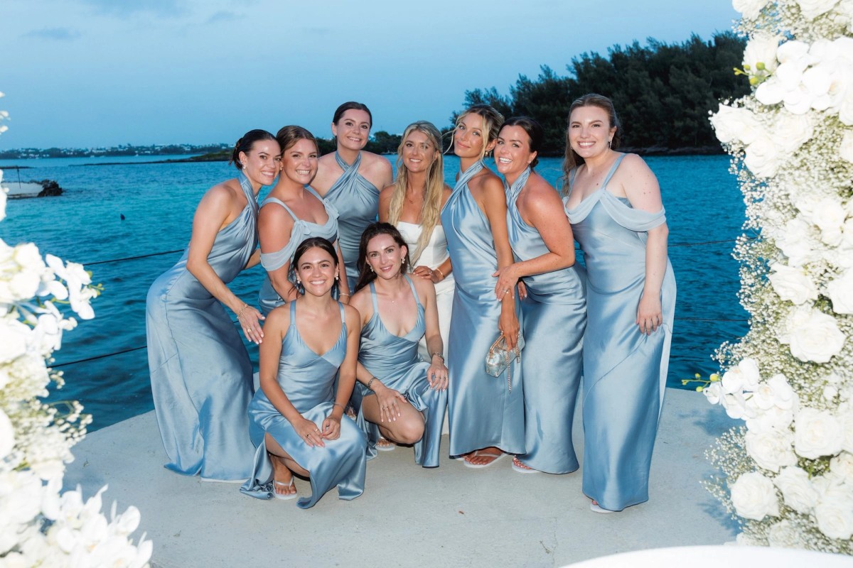 Bridesmaids in blue satin gather at the floral arch by the water on Hawkins Island, Bermuda