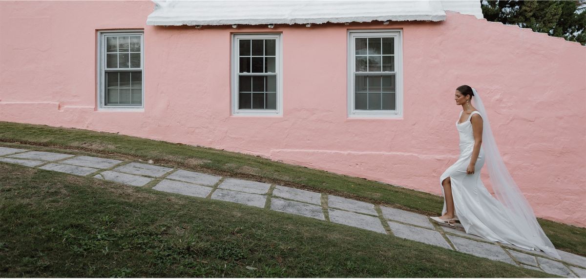 Bride walking a stone path past a pink Bermuda cottage with her veil trailing