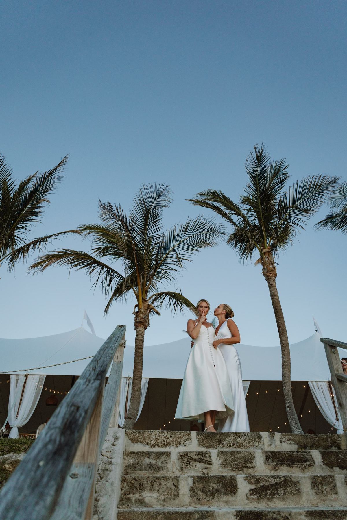 Two brides standing together on a stone wall framed by palm trees in Bermuda