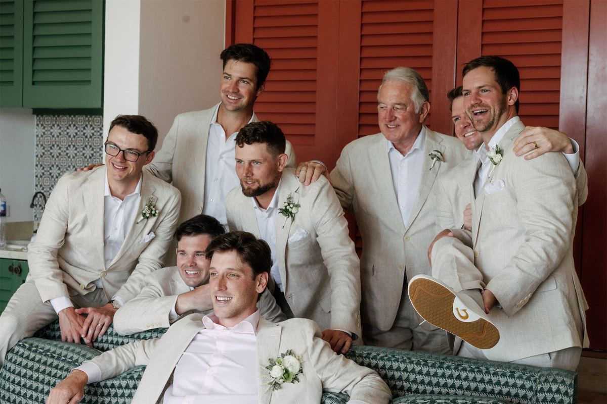 Groomsmen in linen suits piled onto a sofa on a red-shuttered veranda