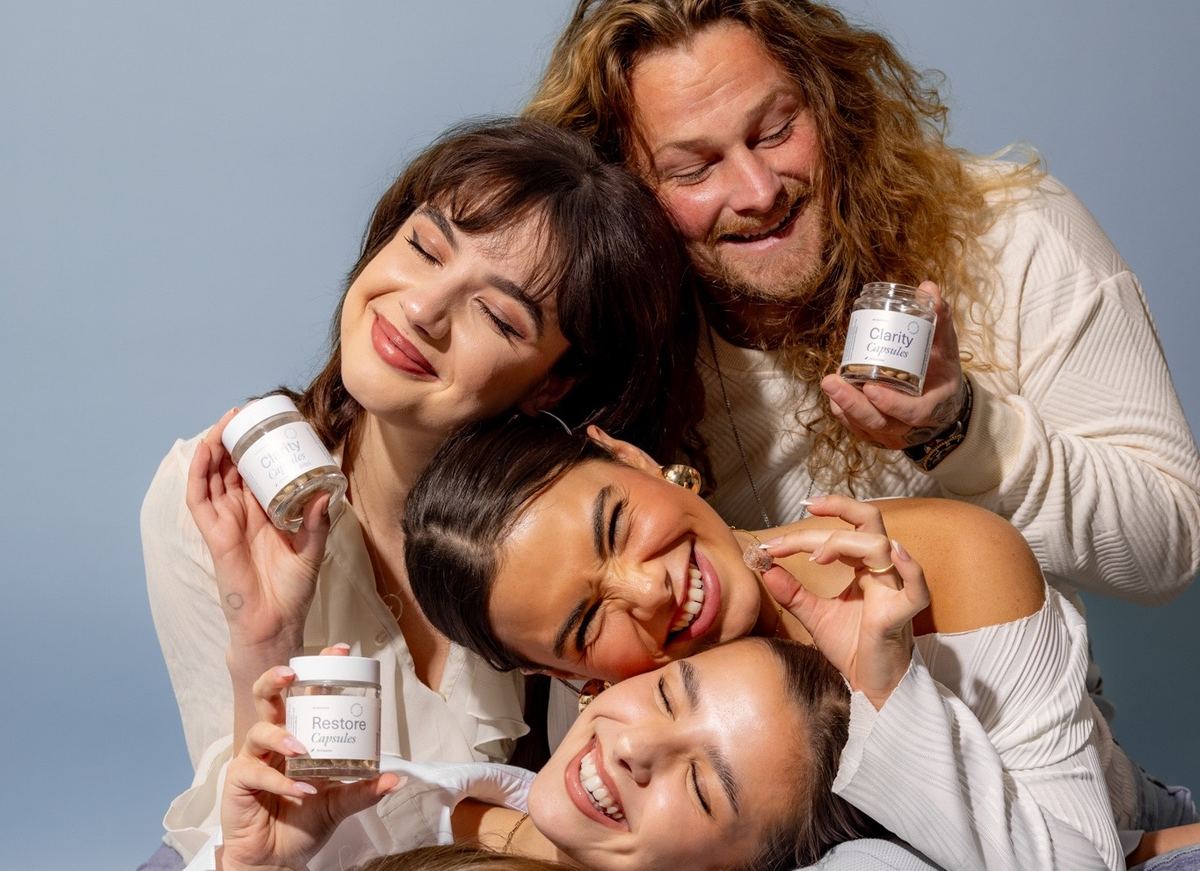 Four models piled together laughing, each holding a jar of capsules
