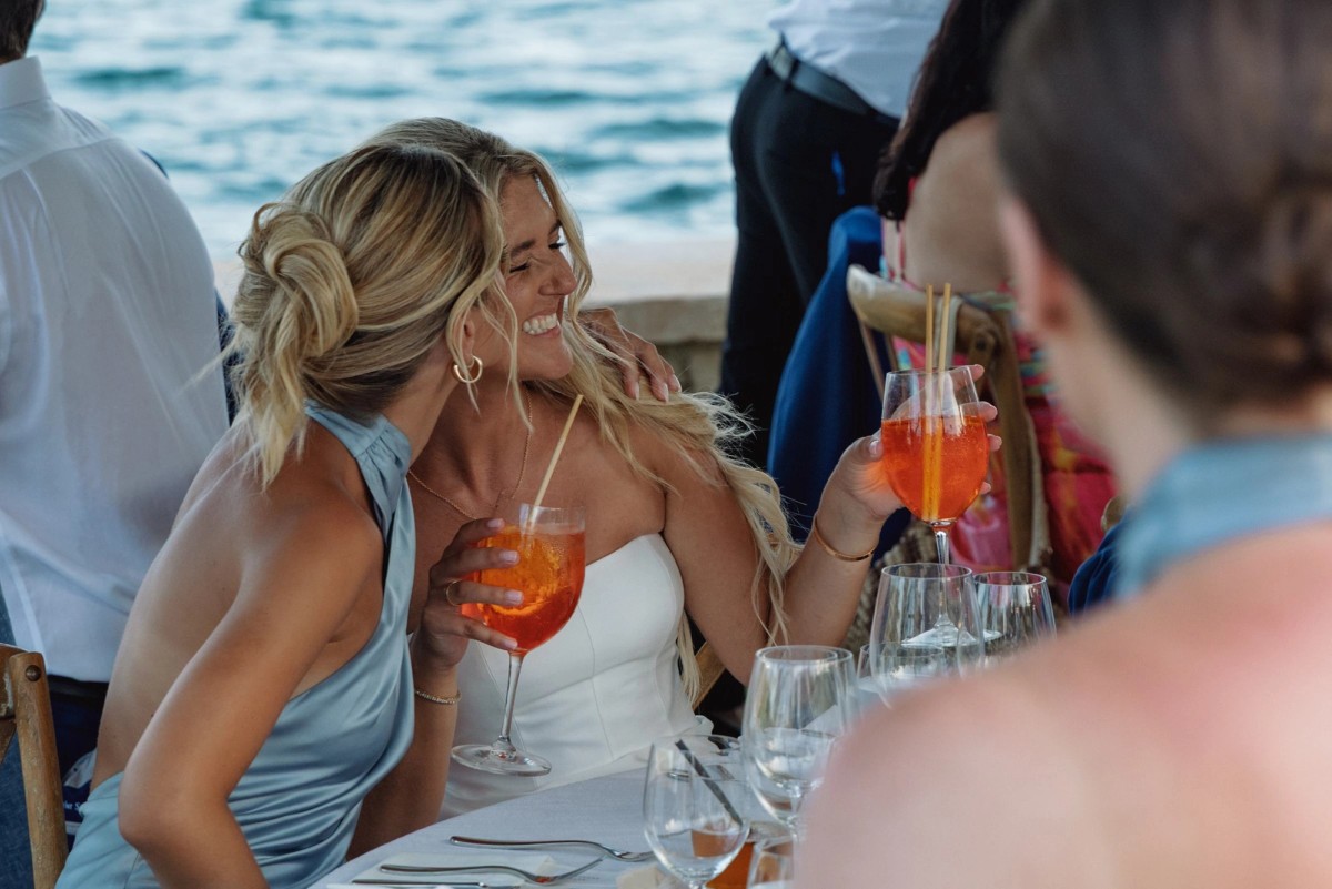 Paige laughs with a bridesmaid over spritzes at the waterfront dinner on Hawkins Island, Bermuda