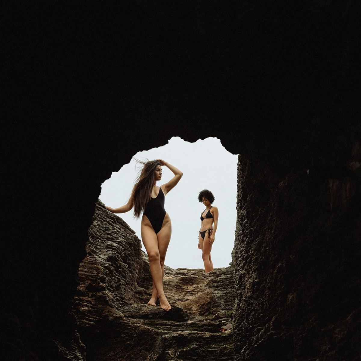 Two swimwear models framed inside the mouth of a dark sea cave