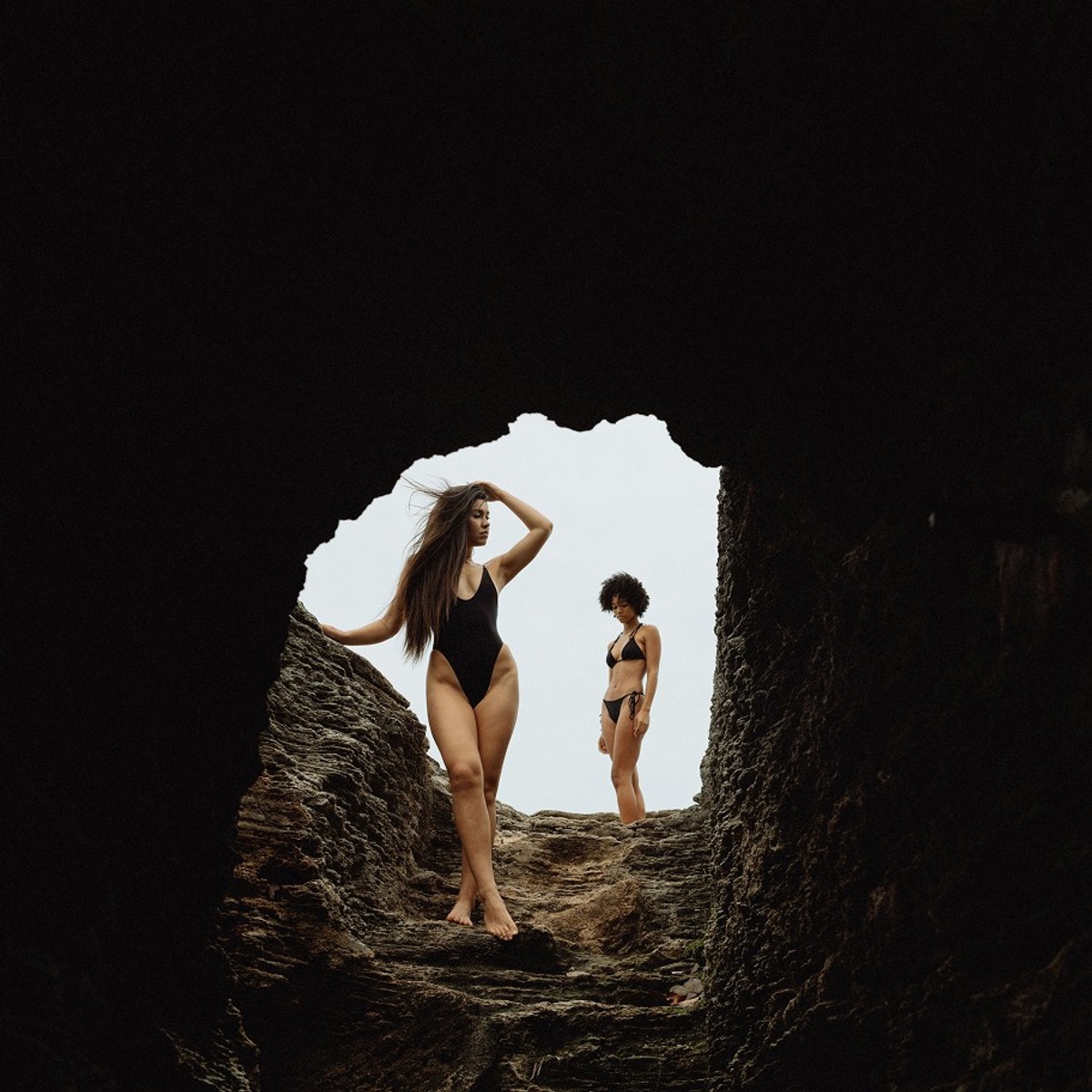 Two swimwear models framed inside the mouth of a dark sea cave