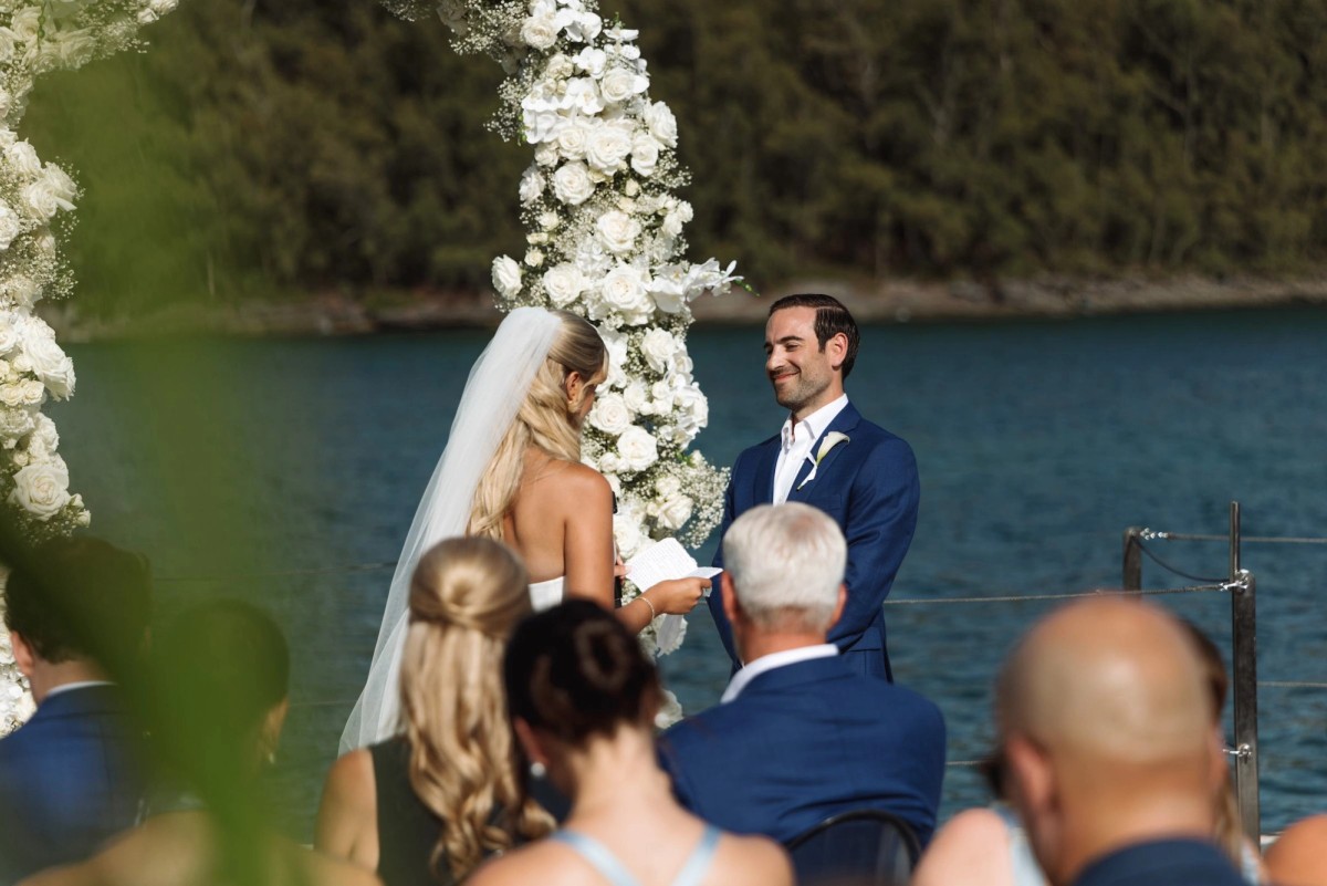 Paige and Cam face each other under the floral arch as guests look on on Hawkins Island, Bermuda