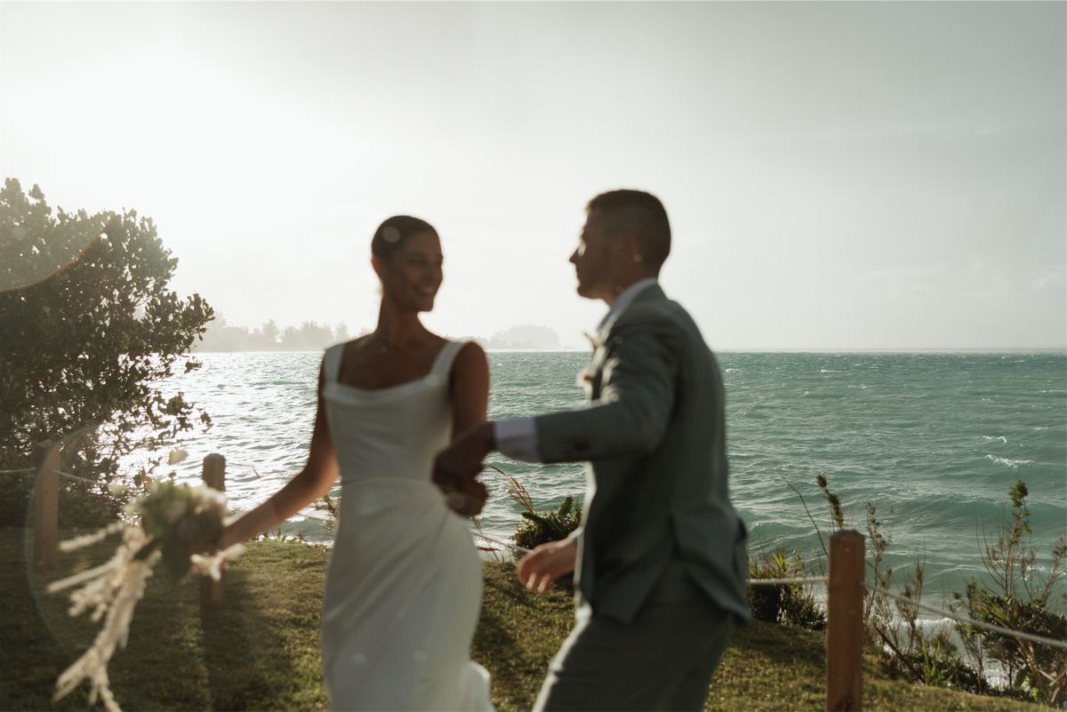 Bride and groom turning together on a bluff above the ocean, bouquet in hand