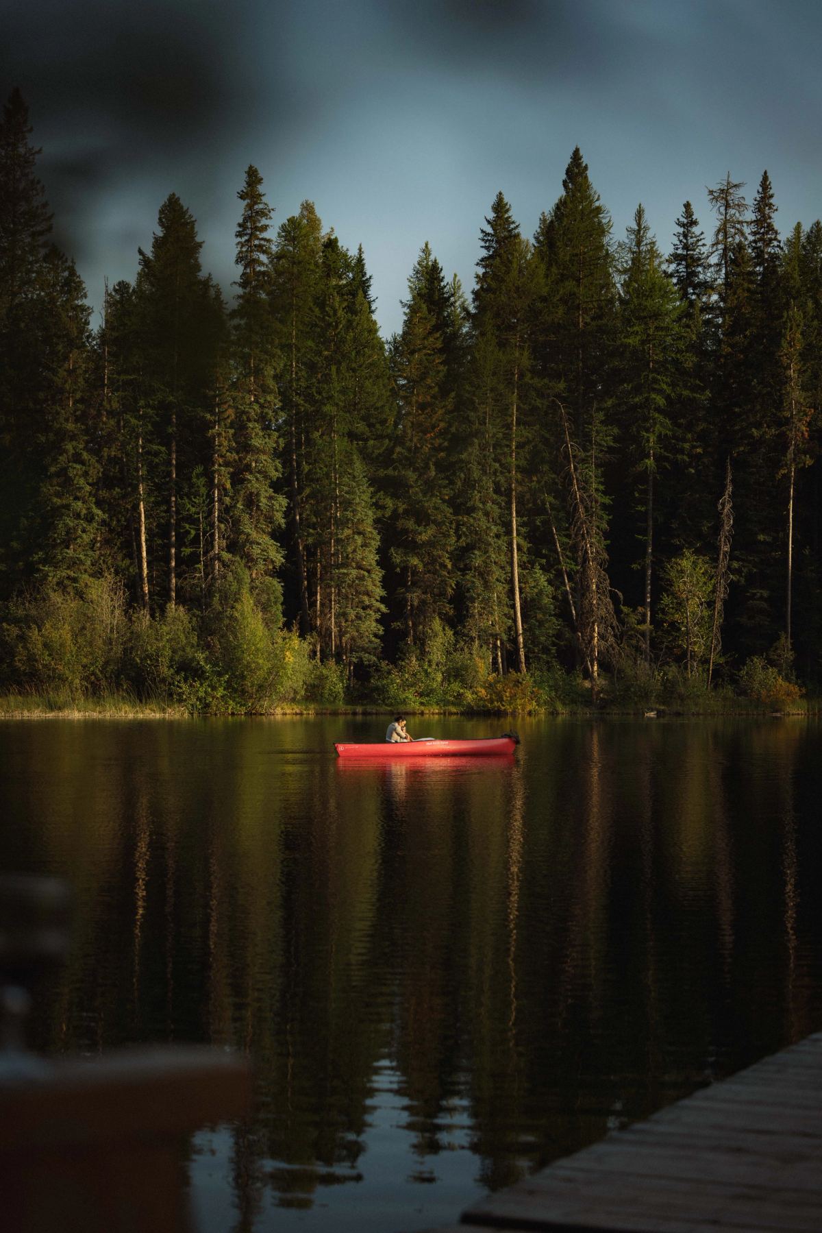 Red canoe crossing a dark forest lake at dusk seen from the dock