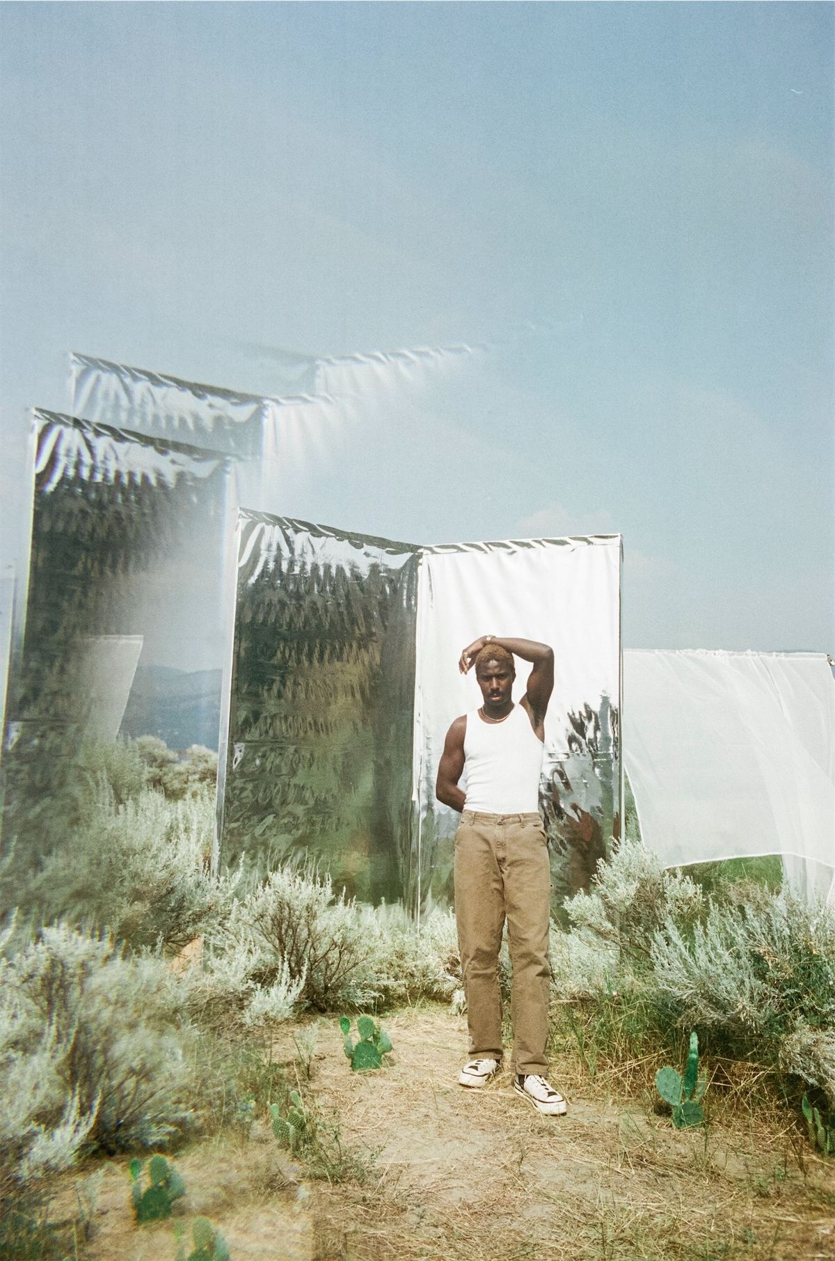 Model standing in desert brush beside a tall mirror reflecting the open sky