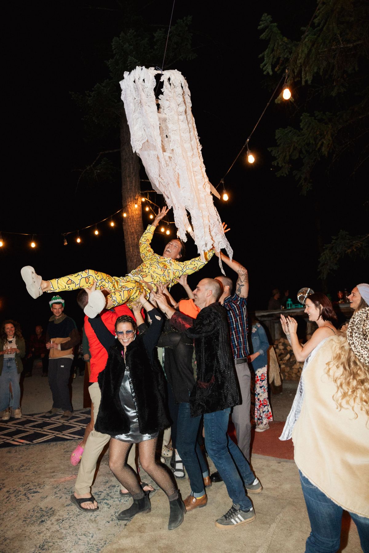 Guests lifting a bride overhead on the dance floor under string lights at night
