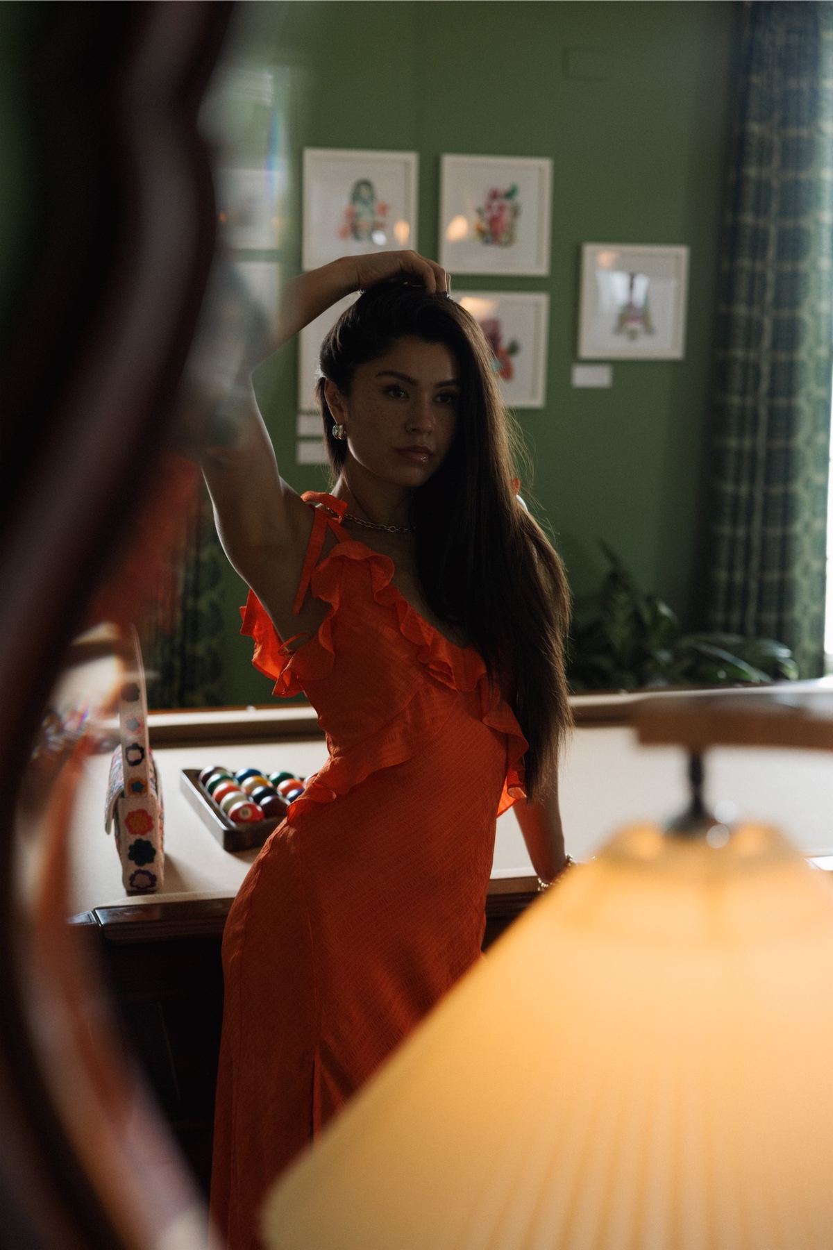 Model in an orange dress posing against a deep green wall beside a lamp