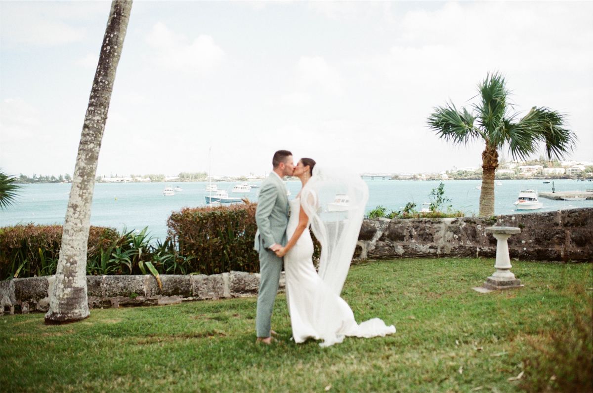 Bride and groom kissing on the lawn framed by palms above the harbour