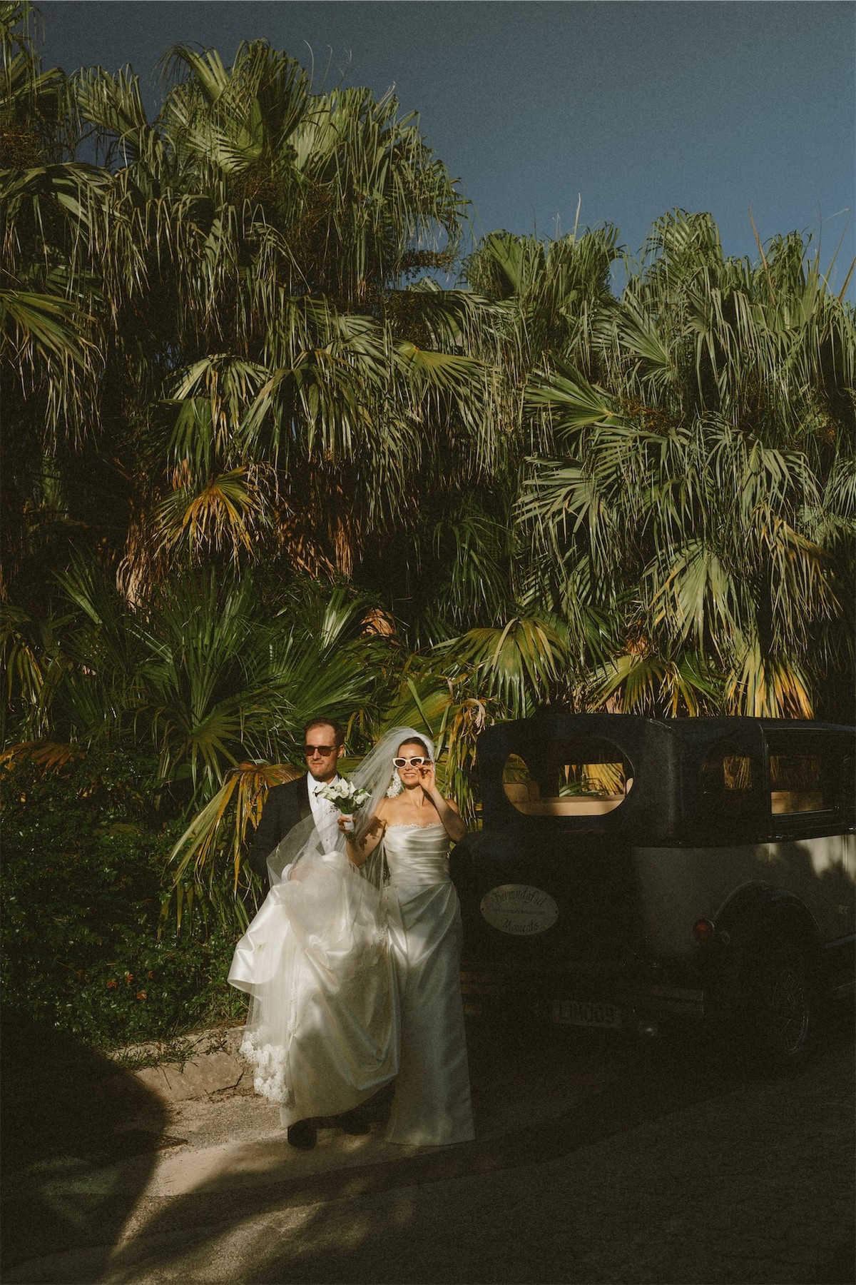 Bride and groom beside a vintage car under a wall of palms