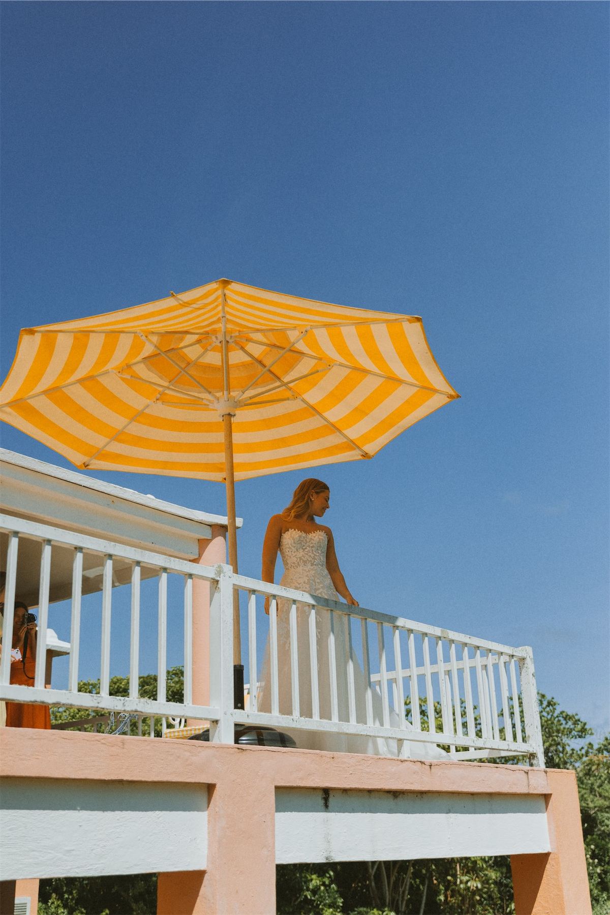 Guest leaning on a balcony rail beneath a yellow striped umbrella against blue sky