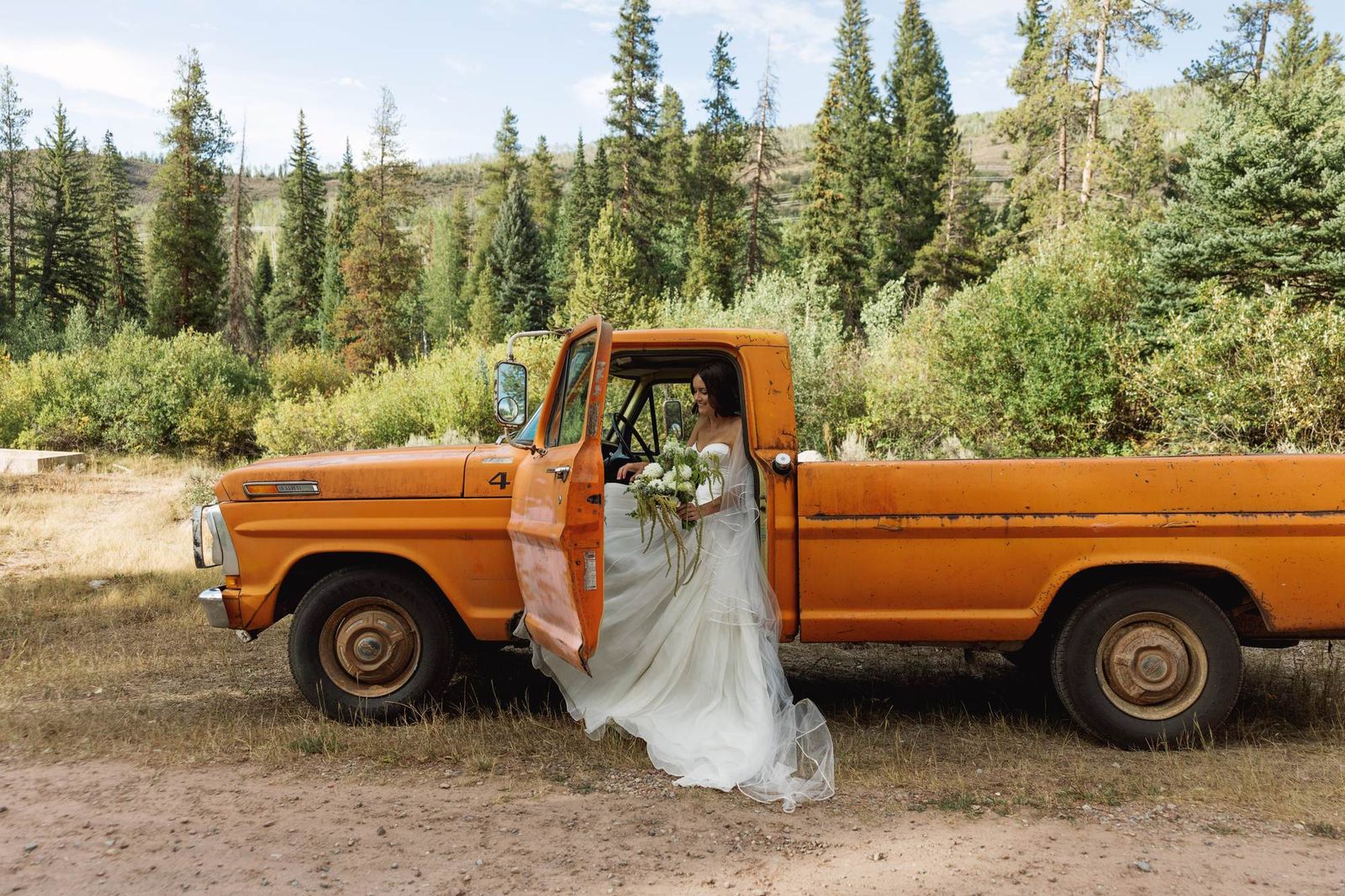 Devin steps out of the orange vintage pickup with her bouquet among the pines at a mountain ranch among the aspens