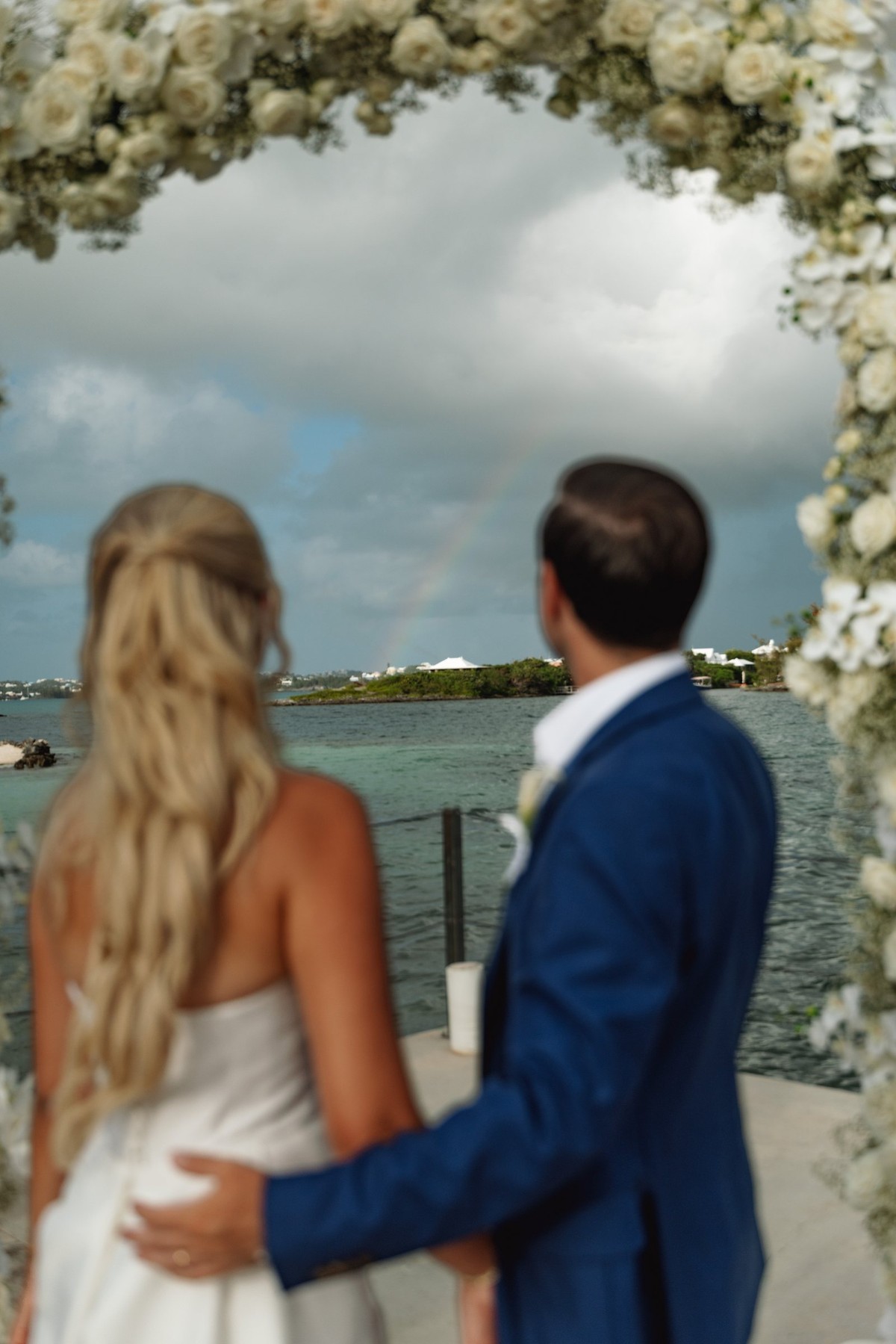 Paige and Cam look out from the floral arch over the bay on Hawkins Island, Bermuda