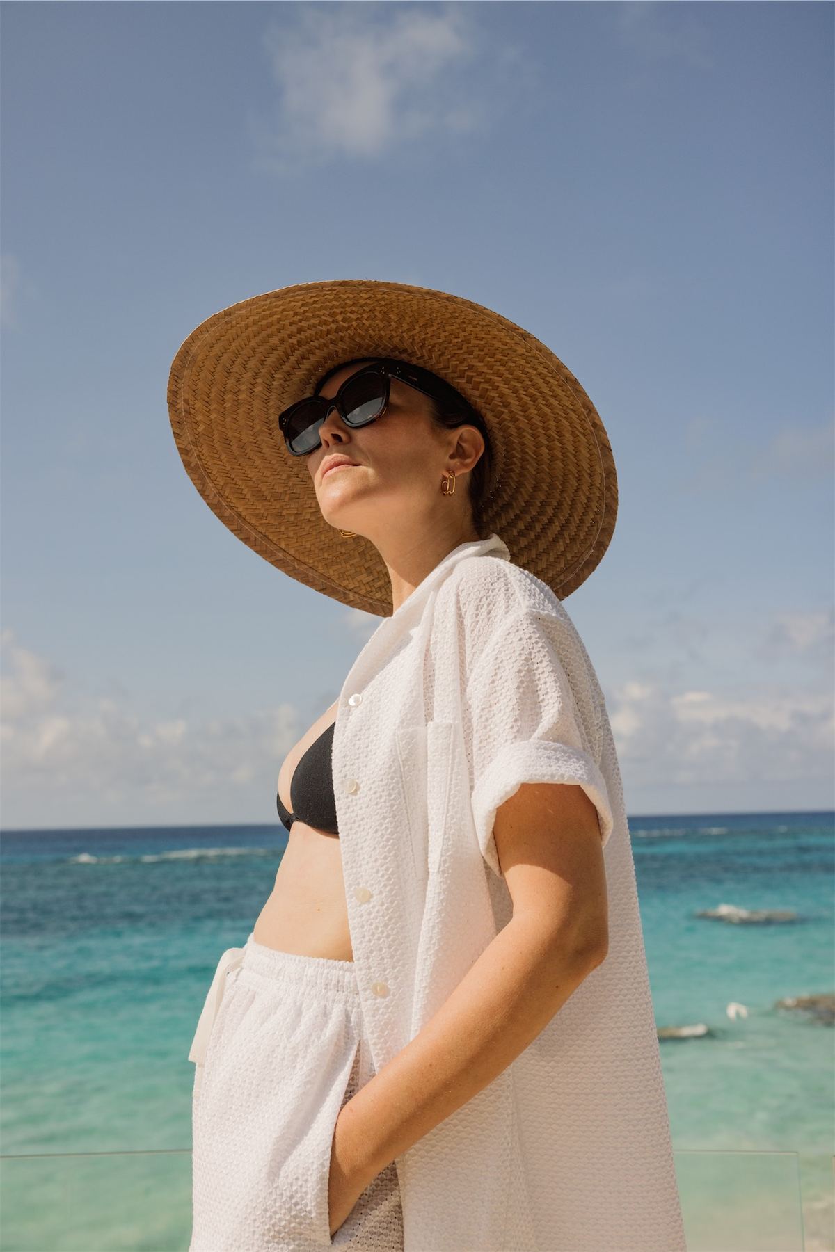 Model in a straw hat and white cover-up tilting her face to the sun on a turquoise beach