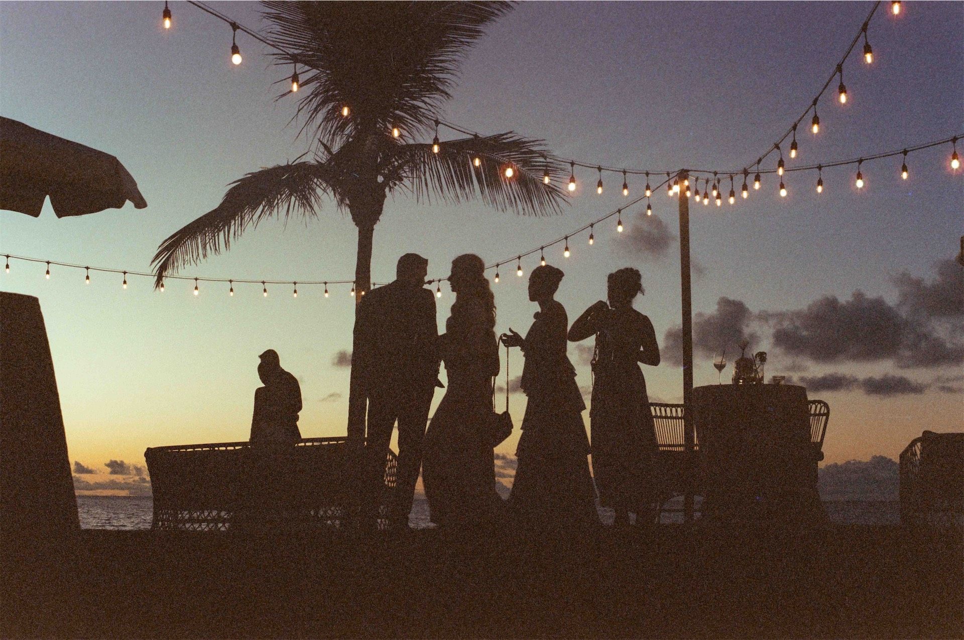 Guests silhouetted against a sunset under strings of festoon lights by the water