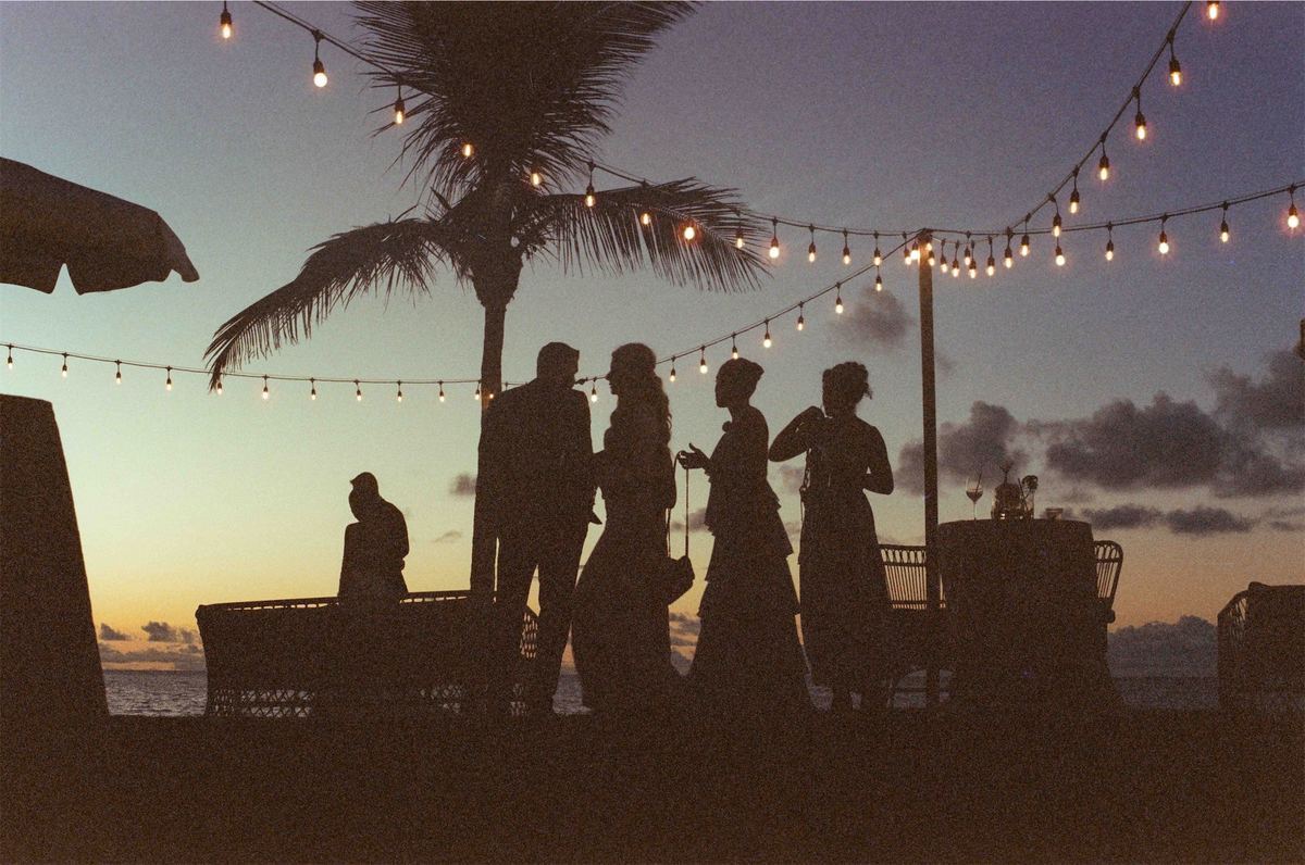 Guests silhouetted against a sunset under strings of festoon lights by the water