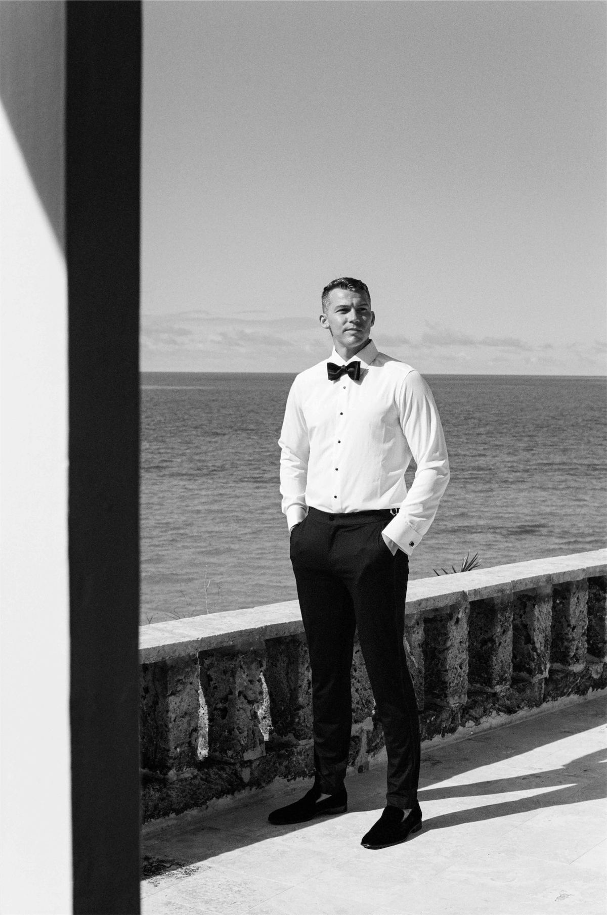 Black and white portrait of the groom standing between columns with the sea behind