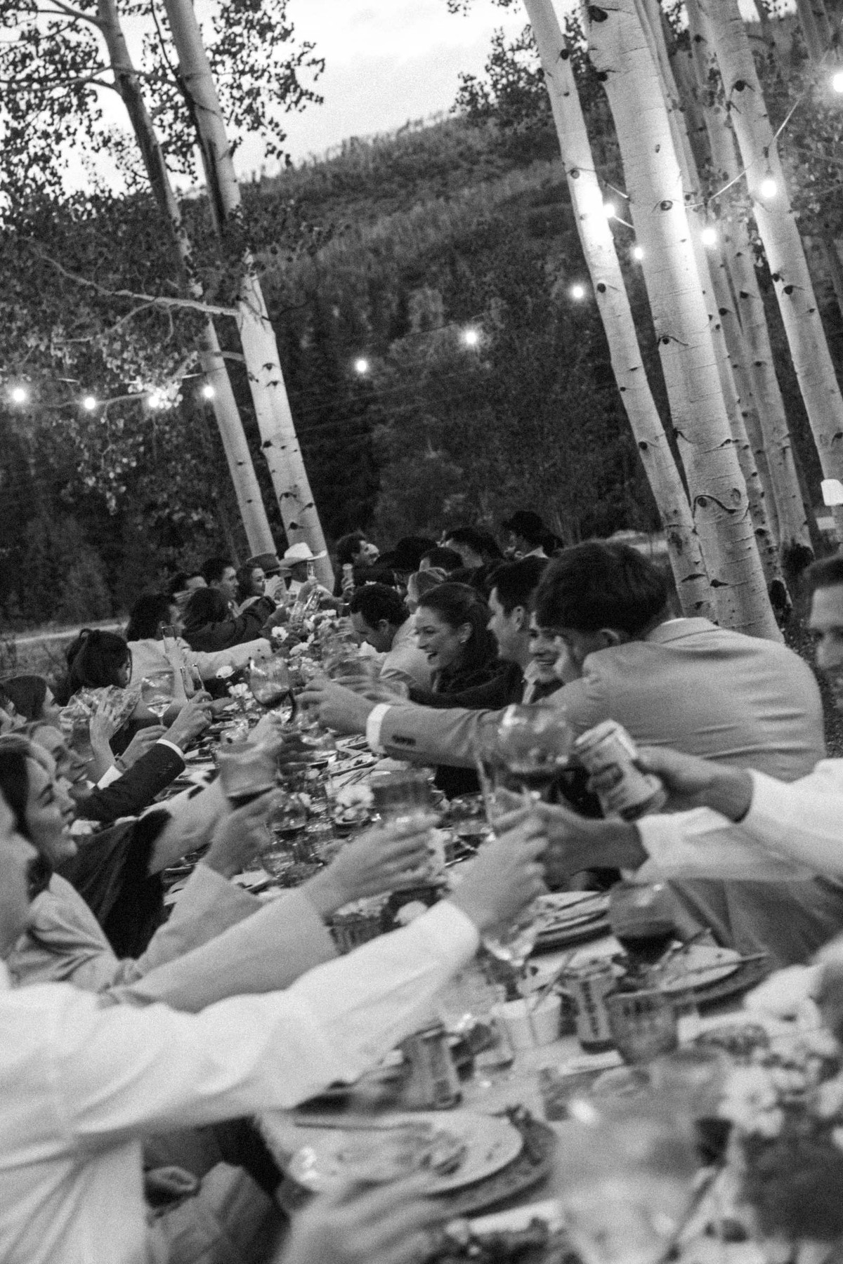 Guests raise their glasses under the string lights at a mountain ranch among the aspens, black and white