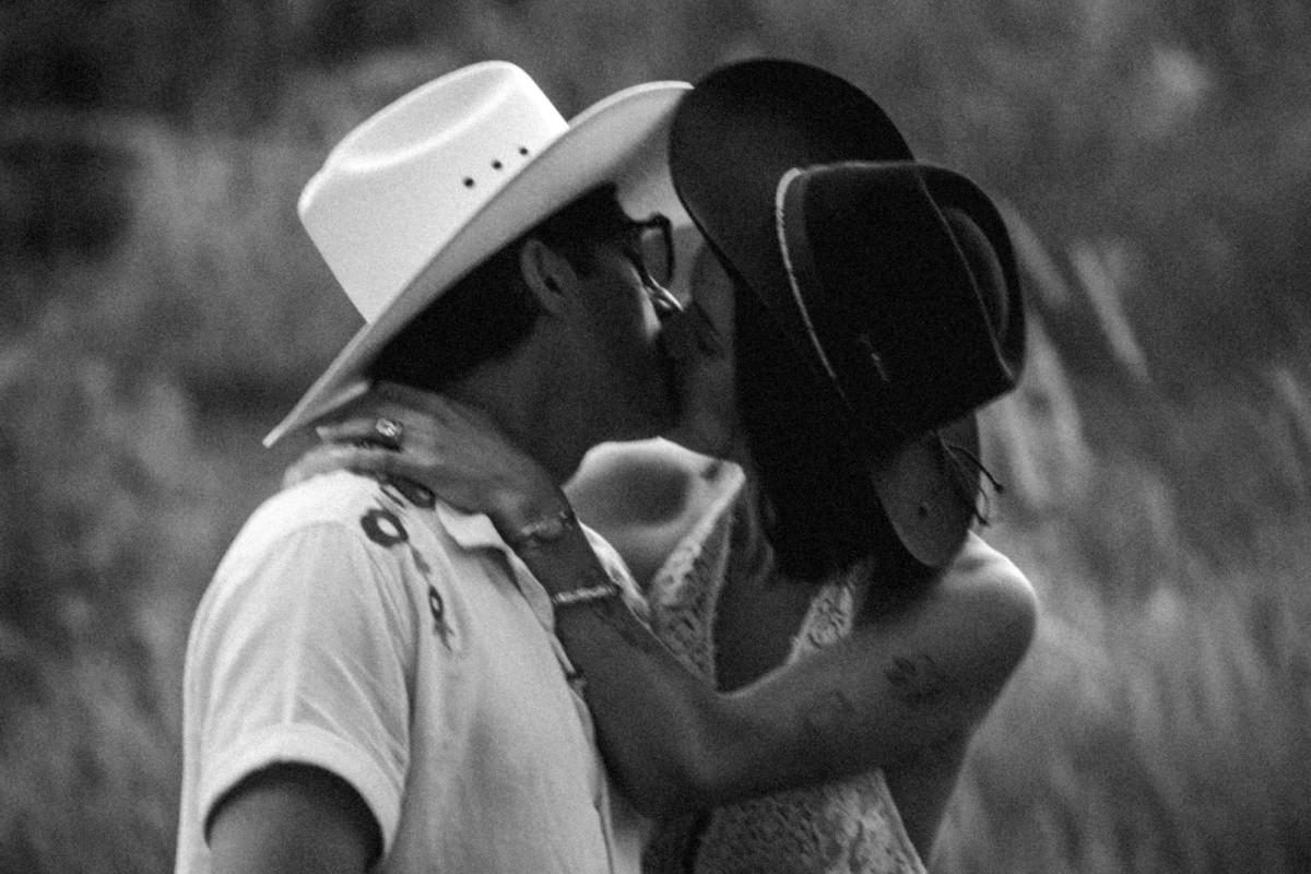 Owen and Devin kiss in their cowboy hats in the tall grass at a mountain ranch among the aspens, black and white