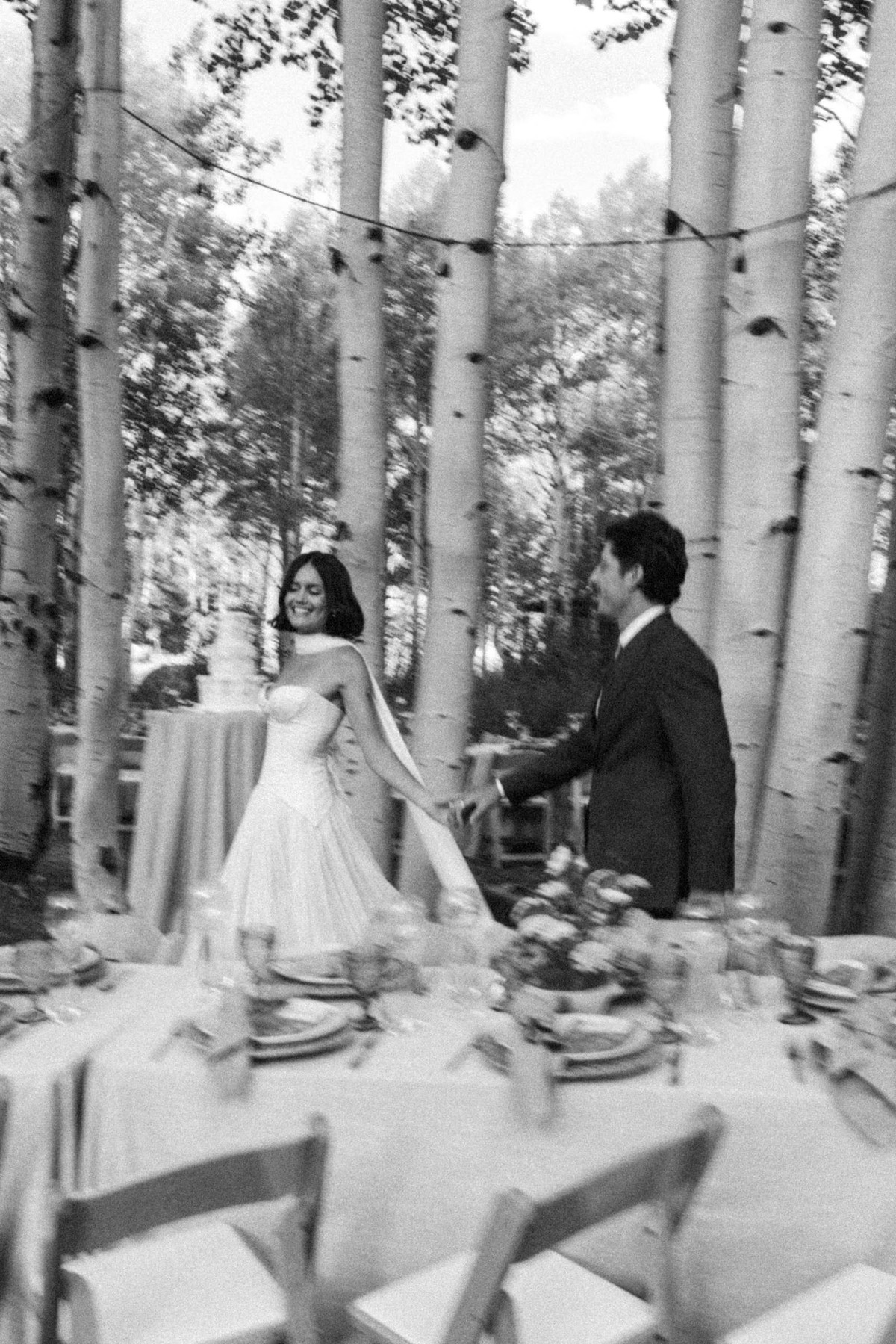 Devin and Owen walk into the reception past the cake at a mountain ranch among the aspens, black and white