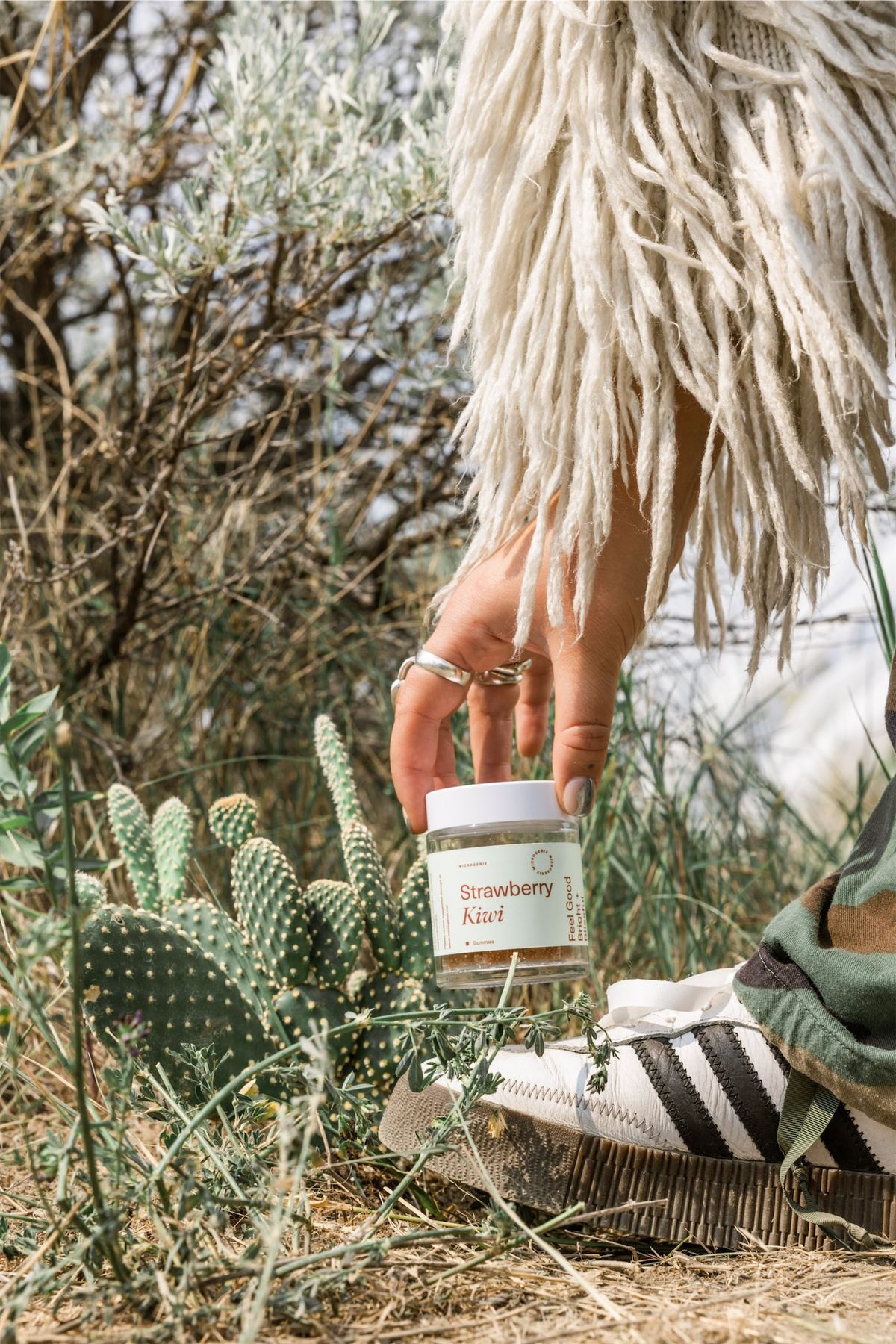 Model in desert scrub holding a Strawberry Kush jar among prickly pear cactus