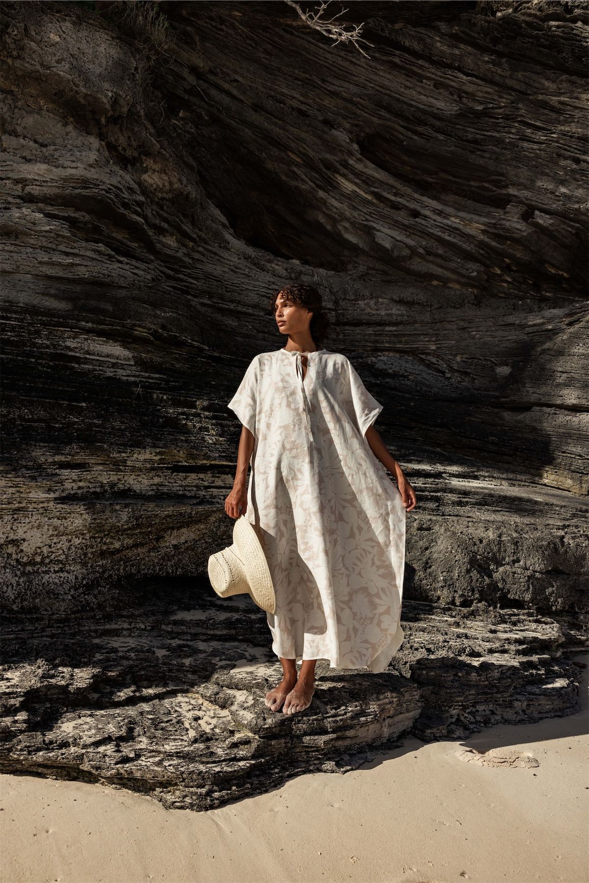 Model in a white kaftan holding a straw hat beside dark driftwood coloured rock