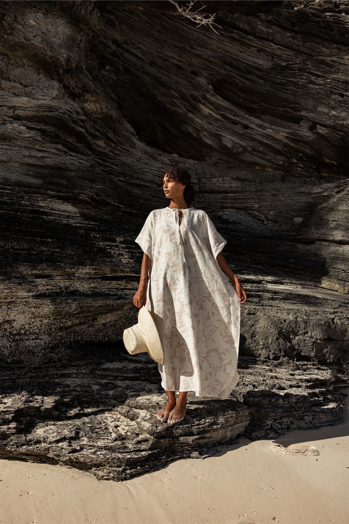 Model in a white kaftan holding a straw hat beside dark driftwood coloured rock
