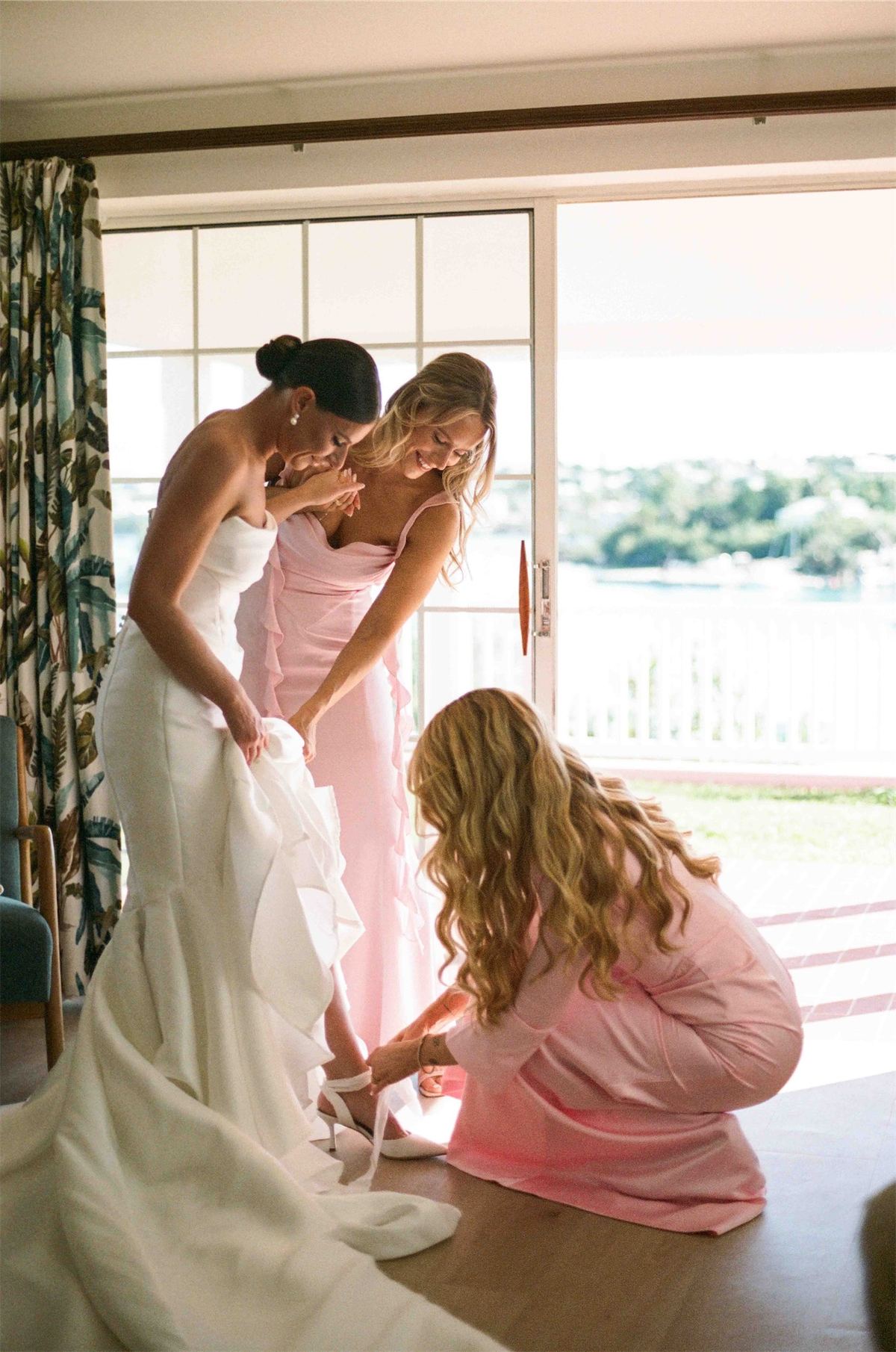 Bridesmaids fastening the bride into her gown beside an open door to the water