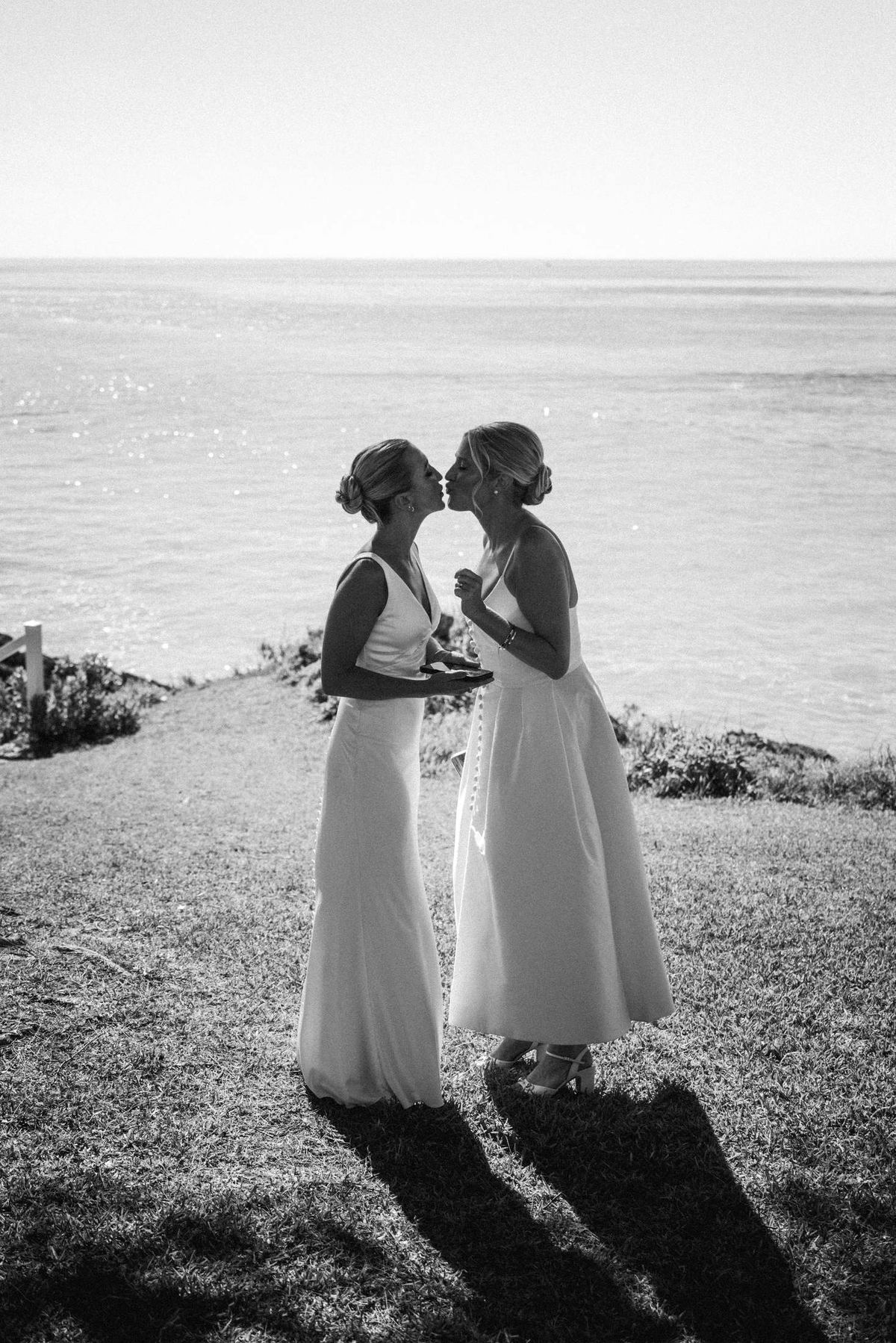 Black and white portrait of two brides forehead to forehead on a Bermuda cliff top with long evening shadows