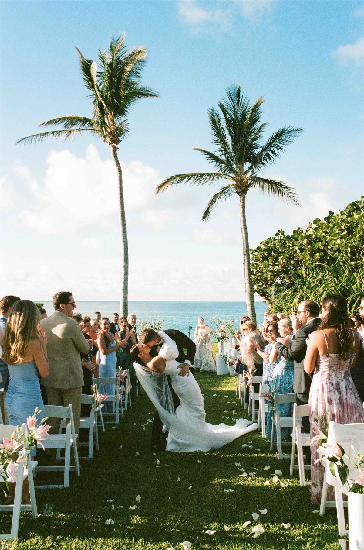 Guests watching the seaside ceremony from white chairs beneath tall palms