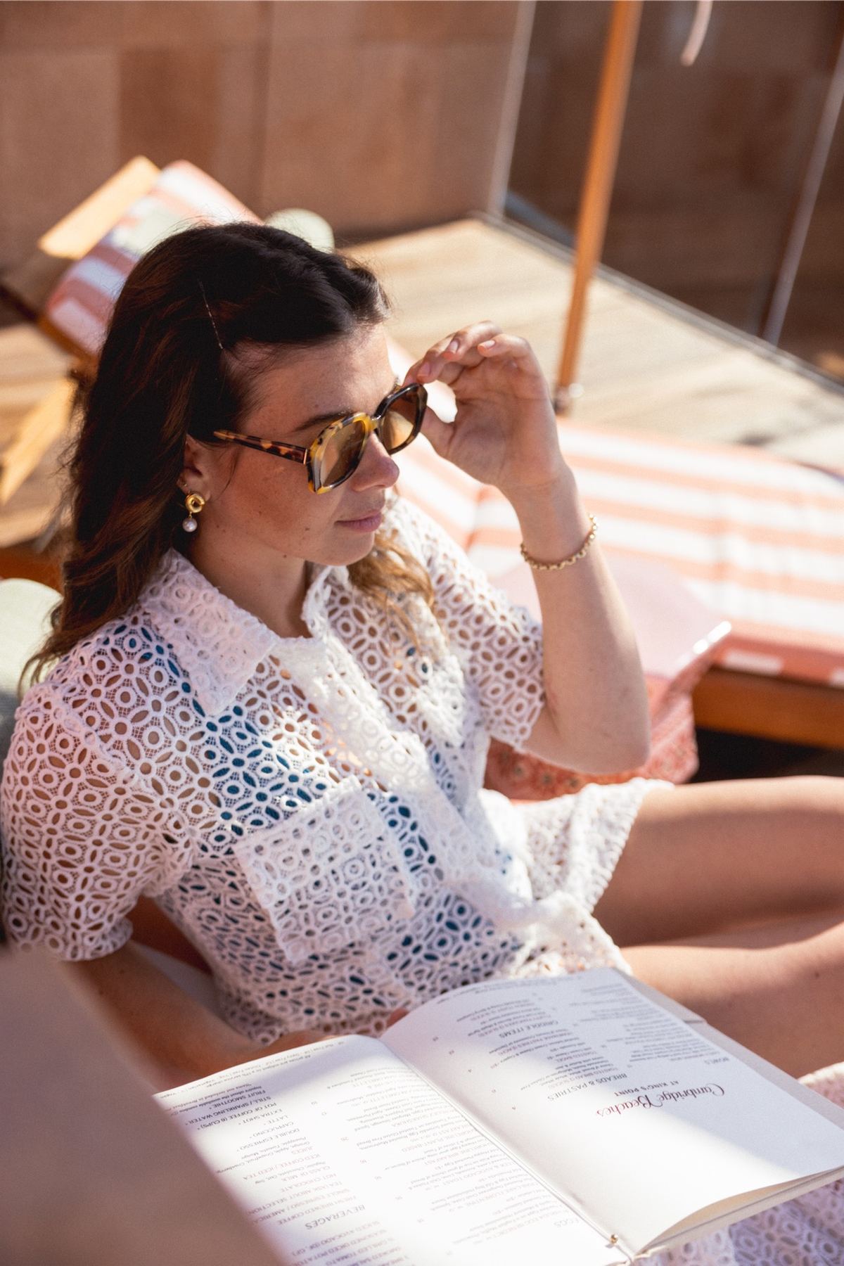 Model reading a magazine in sunglasses and an eyelet cover-up on a striped lounger