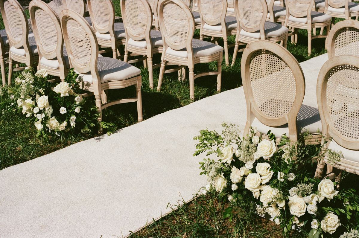 Empty cane ceremony chairs lining an aisle edged with white and green florals