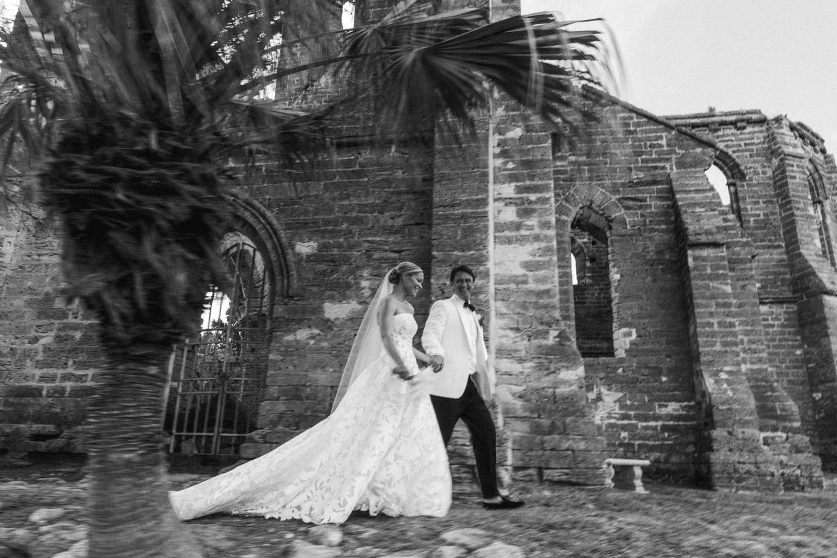 Black and white photograph of the couple walking the Bermuda church ruins through blurred palms