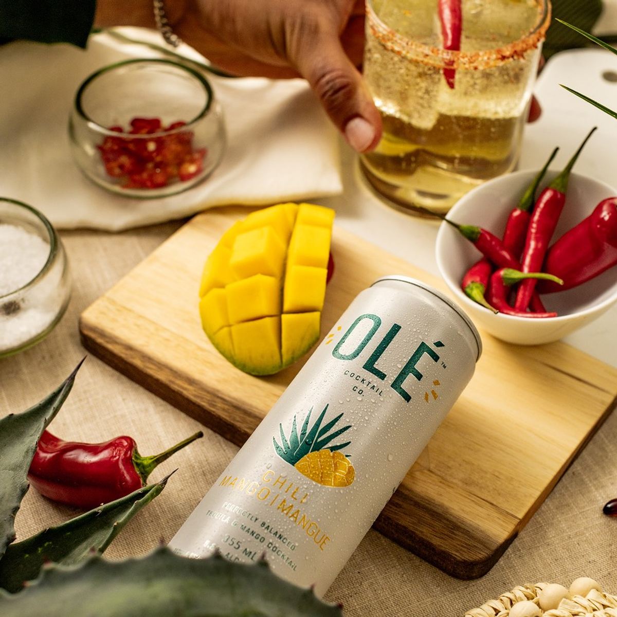 Pineapple cocktail can on a board with mango and chillies as a drink is poured behind