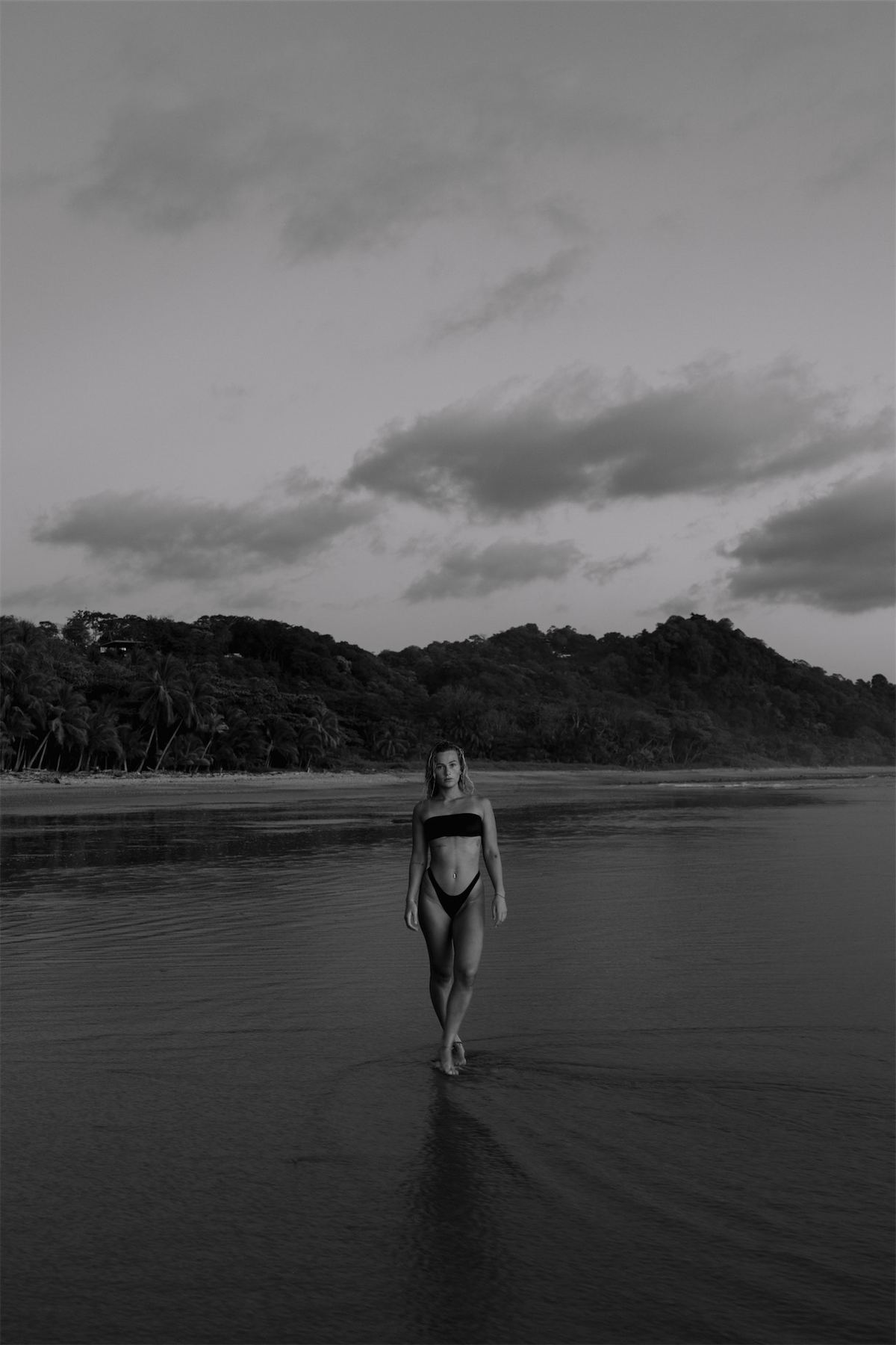 Black and white shot of a swimmer walking into flat water below a forested headland