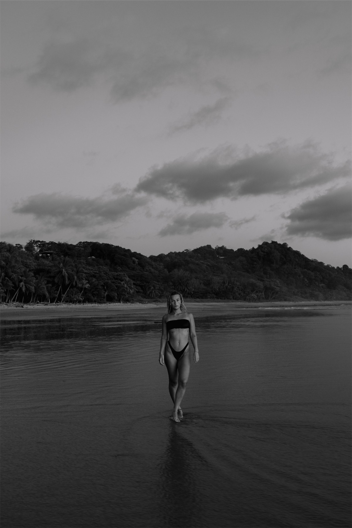 Black and white shot of a swimmer walking into flat water below a forested headland