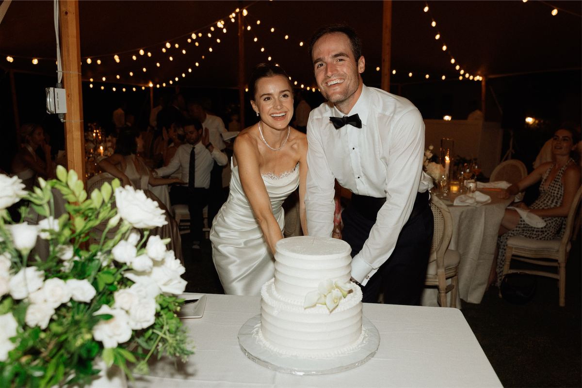Bride and groom cutting a tiered white cake beneath strings of festoon lights