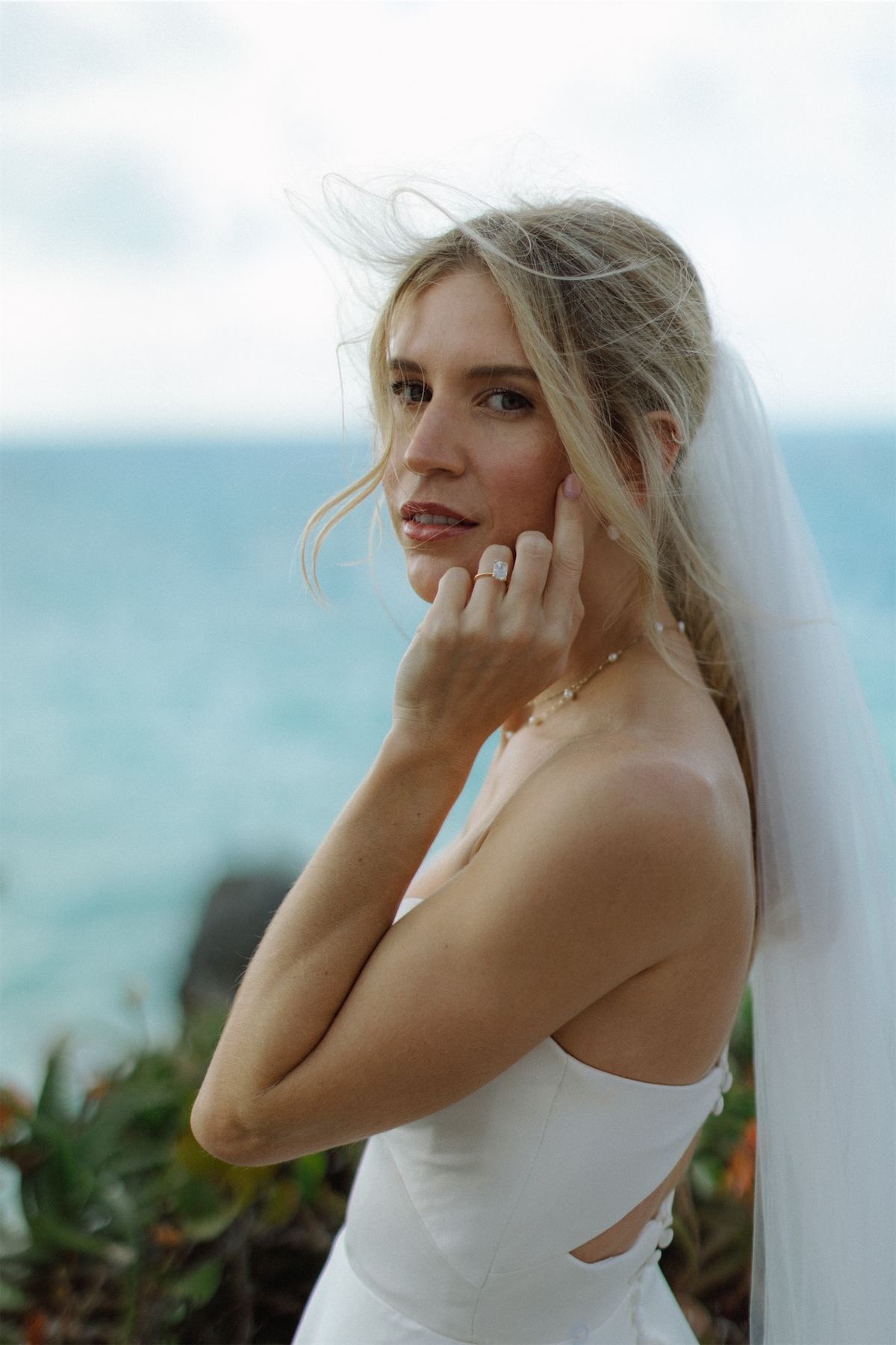 Bride in a long veil glancing over her shoulder with the ocean behind her