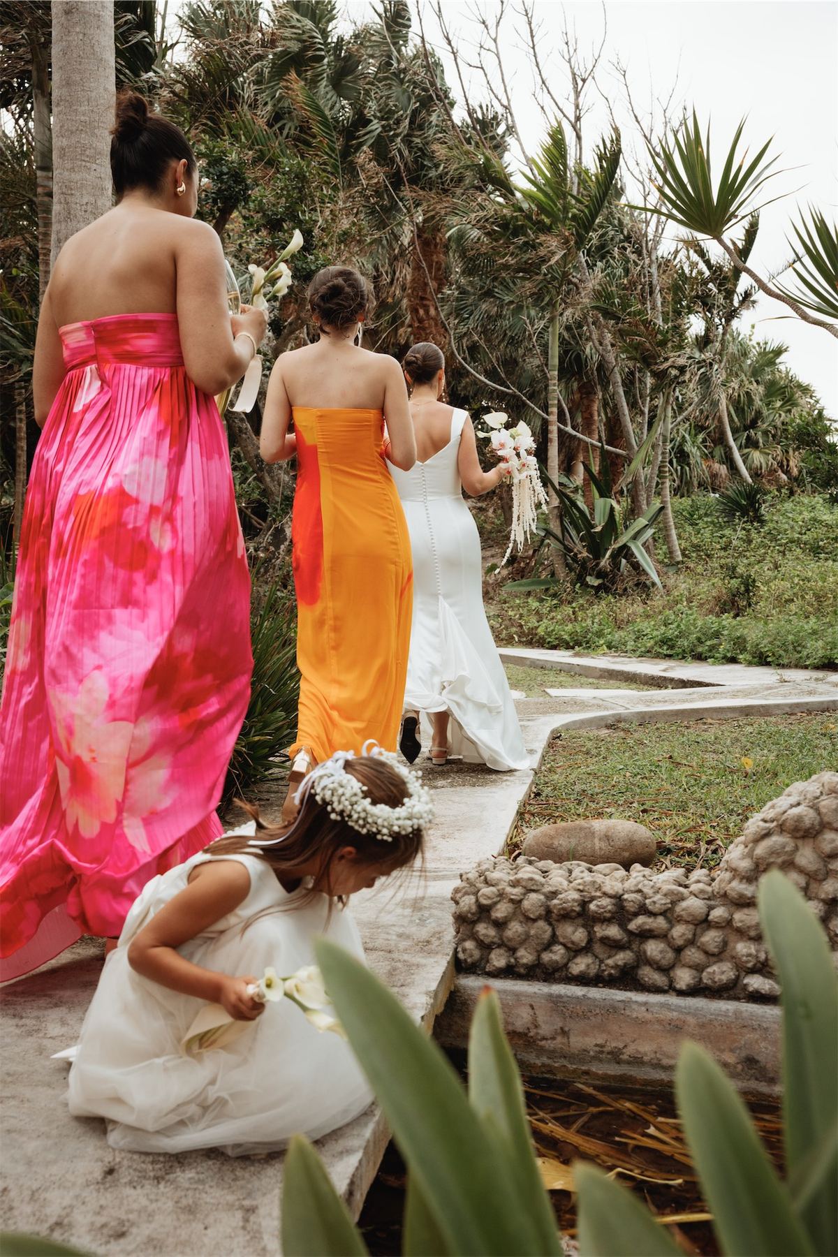 Bridesmaids in bright dresses on a garden path, one helping the flower girl
