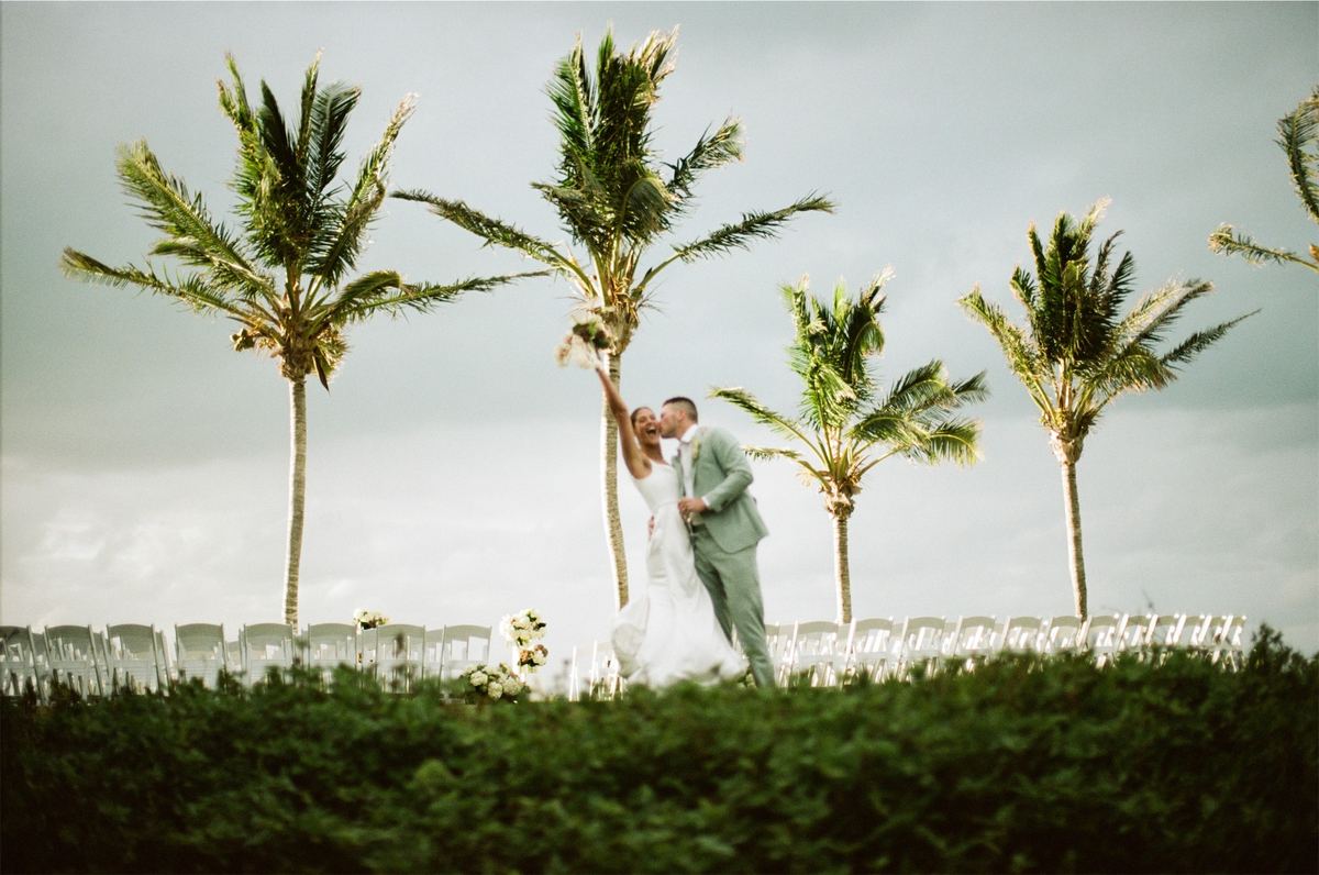 Ceremony framed by tall palms, the couple standing above rows of white chairs by the sea