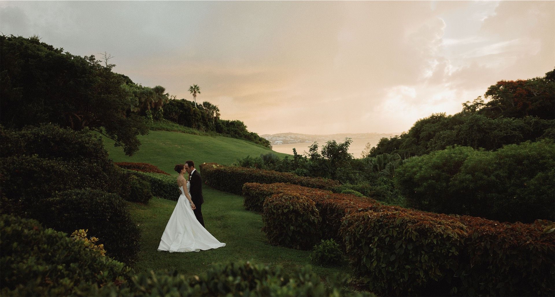 Bride and groom small against a green hillside above the sea at sunset