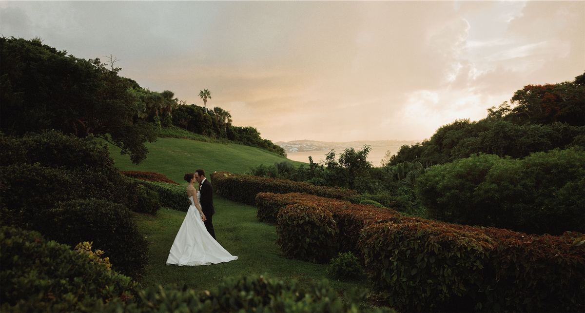 Bride and groom small against a green hillside above the sea at sunset