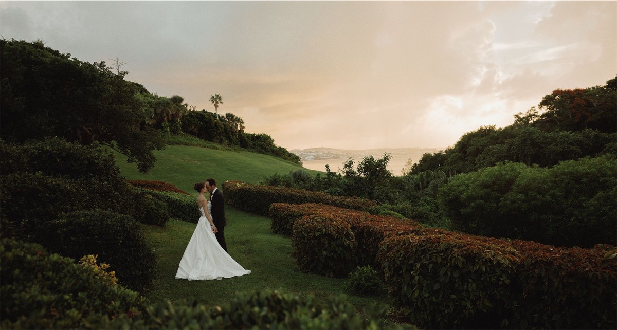 Bride and groom small against a green hillside above the sea at sunset