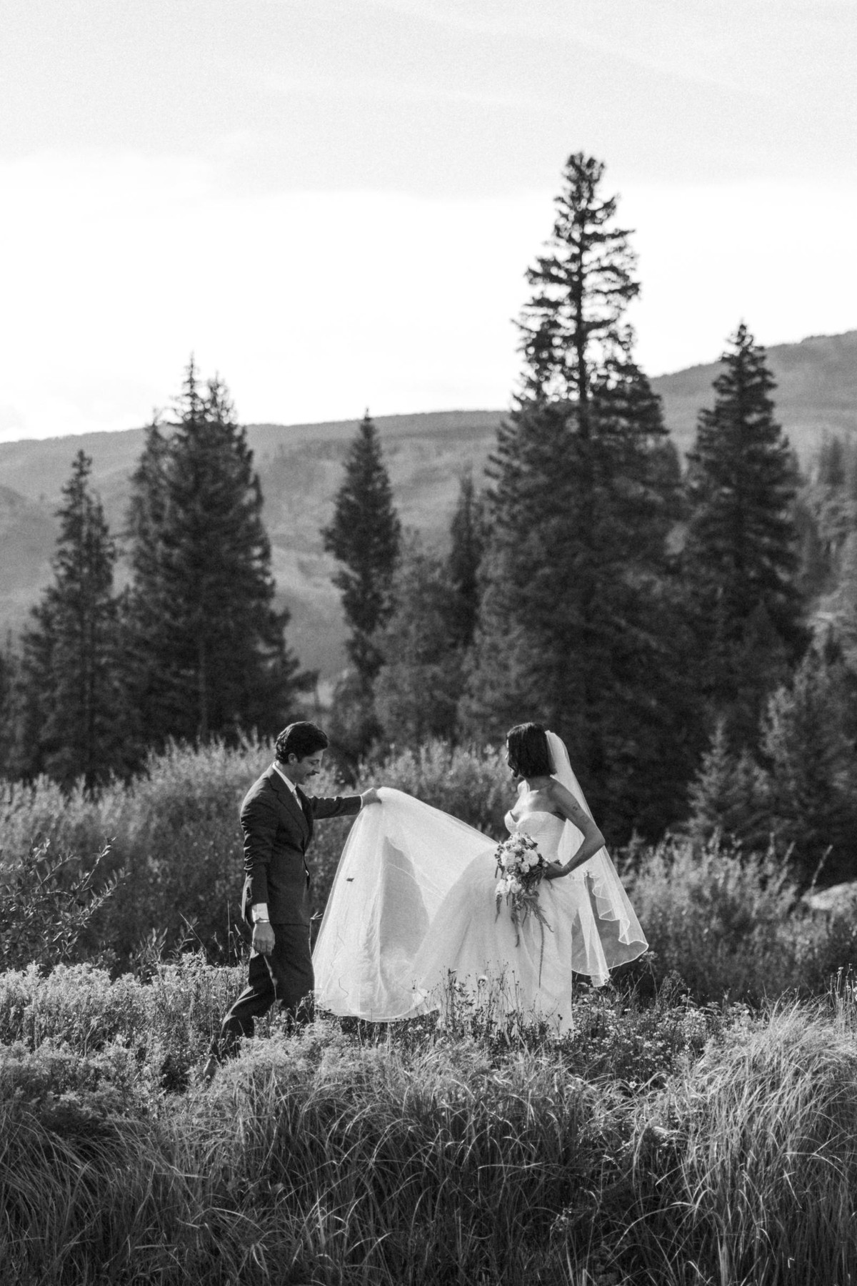 Owen helps Devin with her gown walking through the tall grass at a mountain ranch among the aspens, black and white
