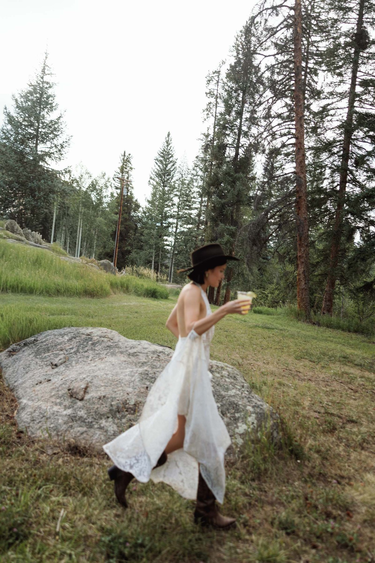 Devin in a white dress, black cowboy hat and boots crossing the meadow with a drink at a mountain ranch among the aspens