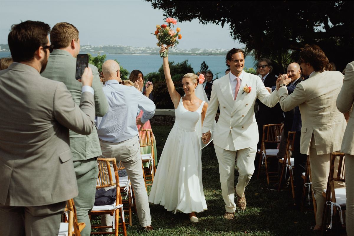 Newlyweds cheering with the bouquet raised as they exit the seaside aisle