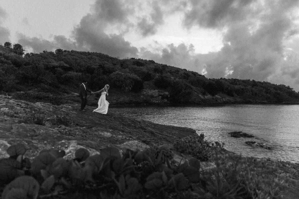 Paige and Cam hand in hand on the rocky shoreline under a stormy evening sky on Hawkins Island, Bermuda, black and white