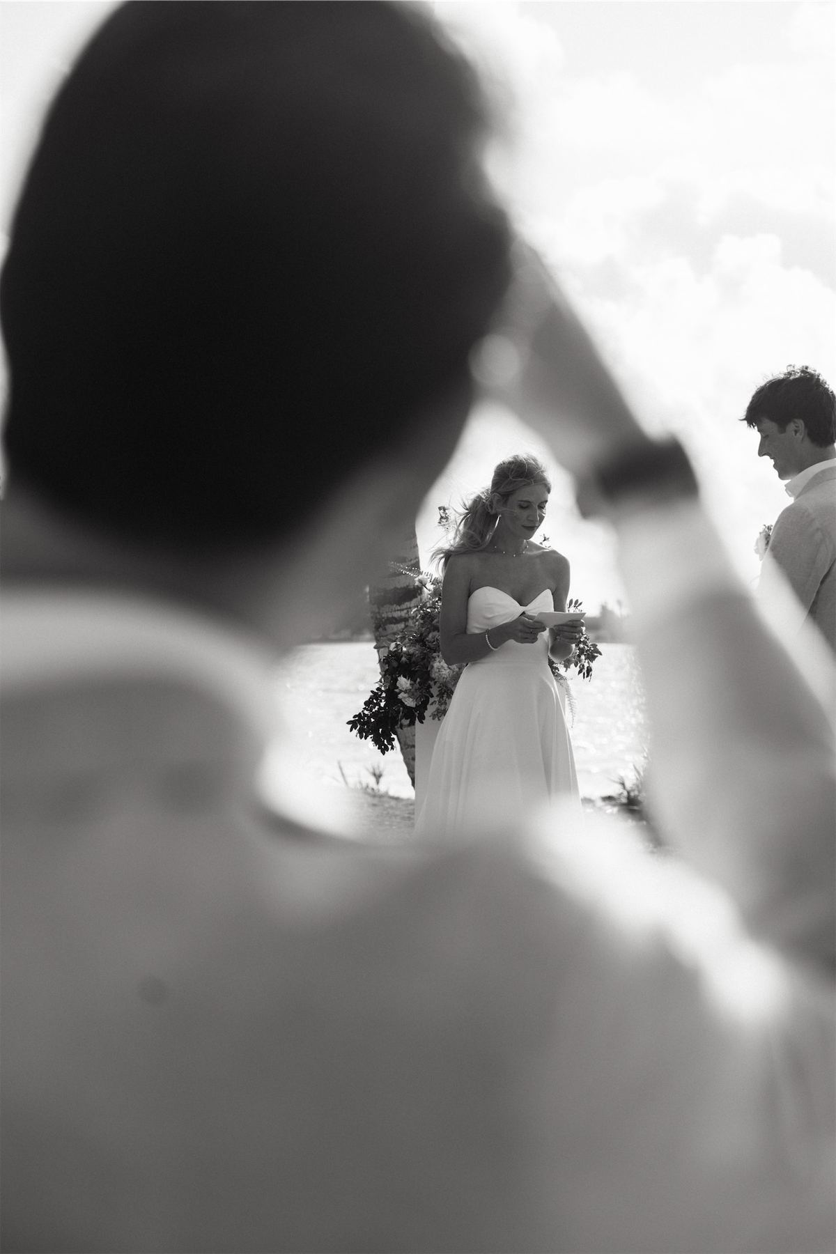 Black and white frame shot past a guest toward the bride during the ceremony